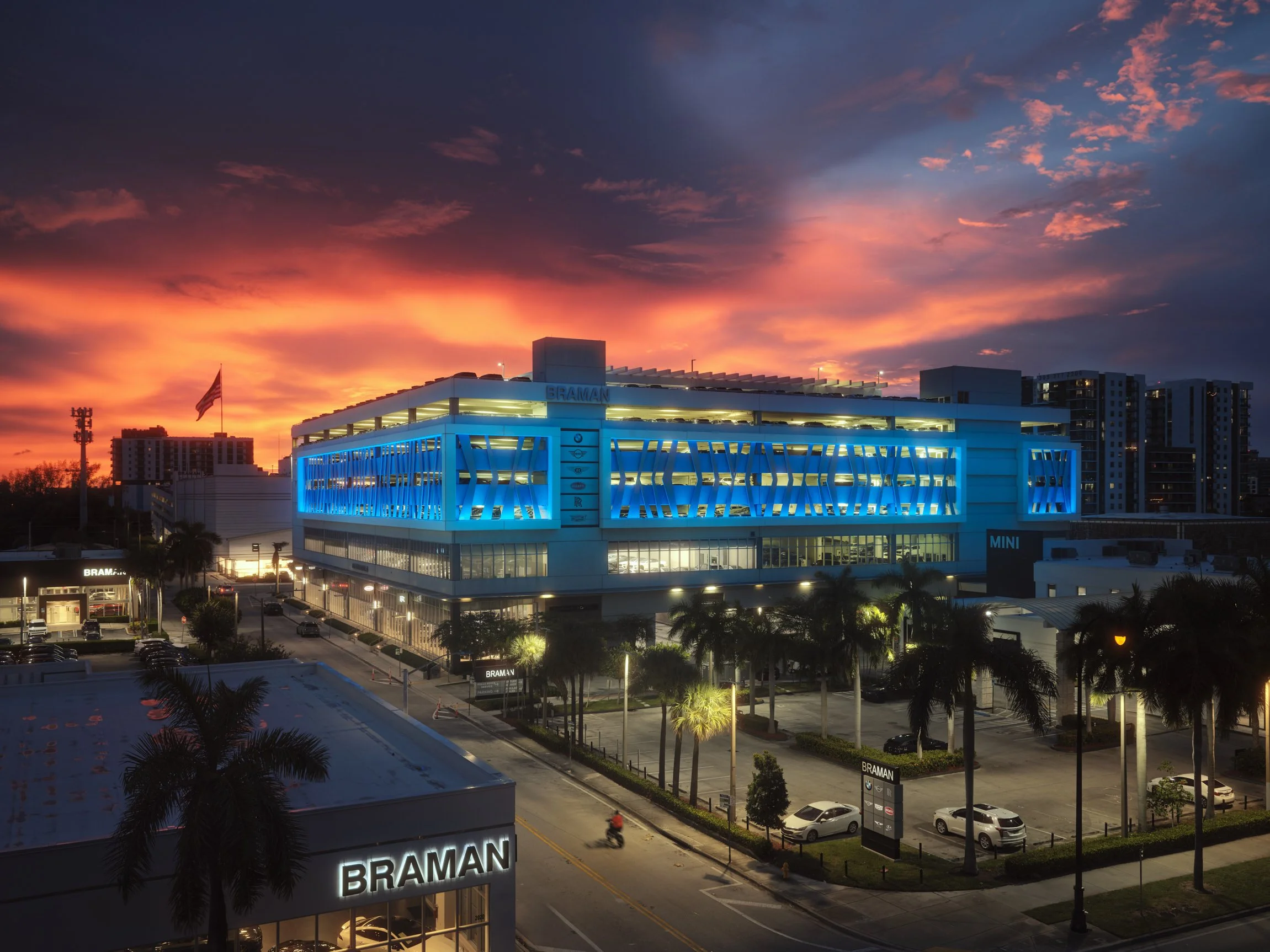 An architectural photograph of the Braman auto showroom & garage in Miami's Edgewater neighborhood at dusk. An intense, beautiful orange sunset contrasts with the blue led light fixtures emanating from the garage's facade. Captured by Miami and Los A