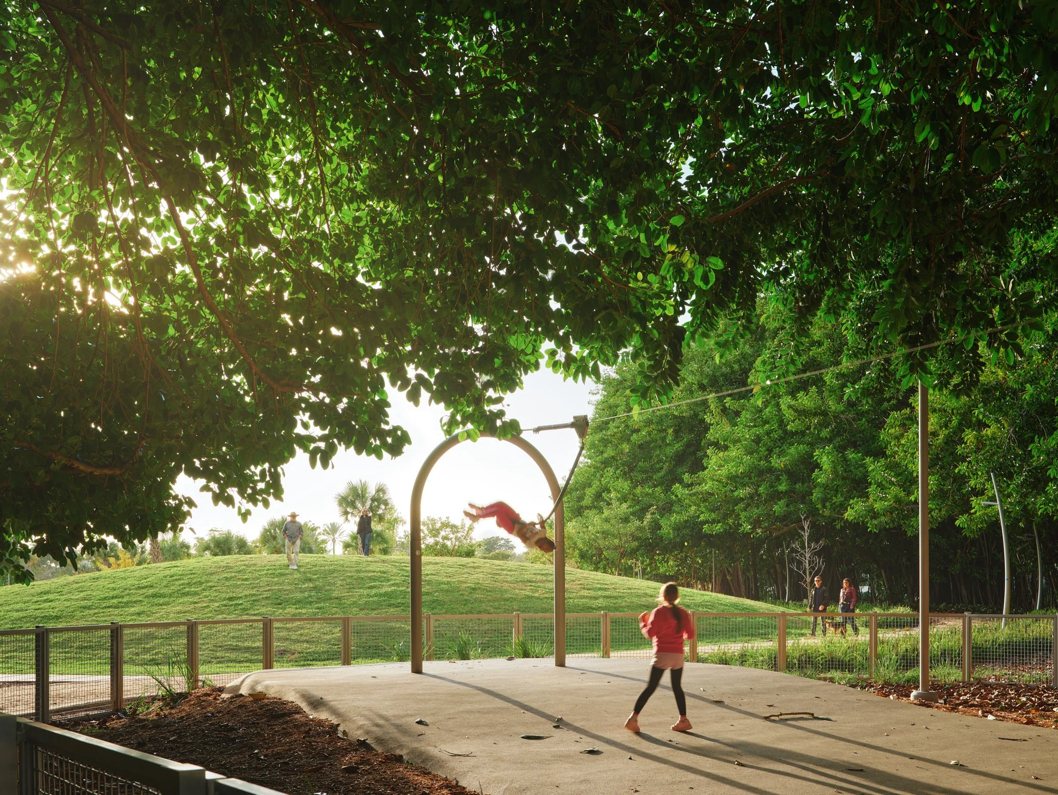 Architectural photograph of a child mid-flight on a zip line beneath the canopy of banyan trees at golden hour in Bayshore Park Miami Beach. Captured by Miami Architectural Photographer Christian Santiago