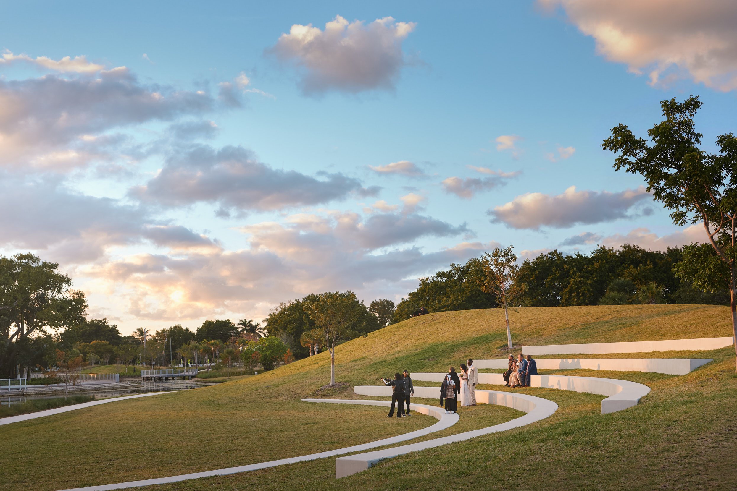 A landscape architectural photograph of a wedding party snapping selfies on the amphitheater at Miami Beach's Bayshore Park. Late golden hour light bathes the scene in soft warm shadows. Captured by Miami Architectural Photographer Christian Santiago