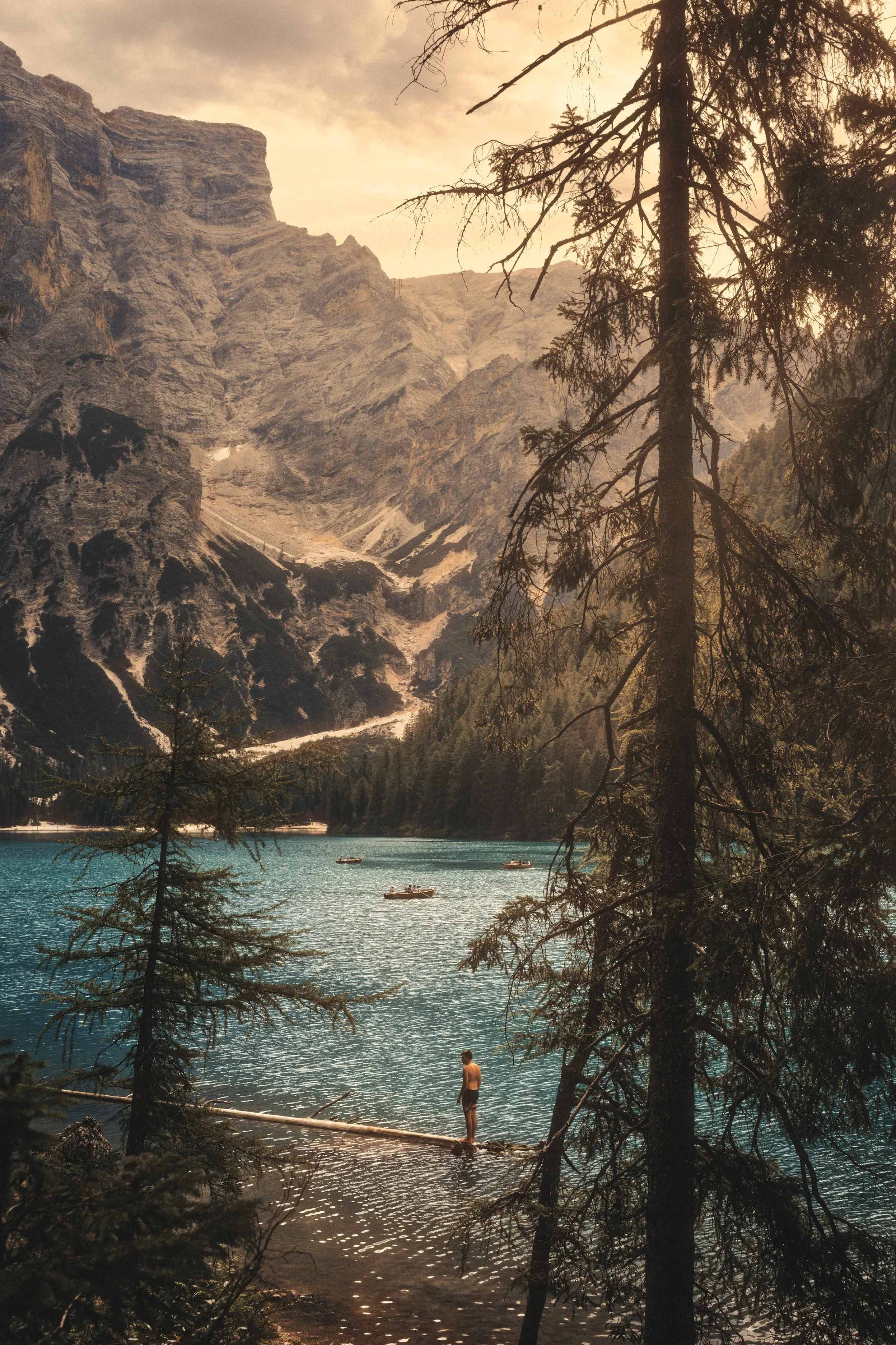 Architectural-style travel photograph of Lake Braies in the Dolomites of northern Italy, featuring turquoise alpine waters, dramatic mountain peaks, and a lone figure standing on a log in the foreground. The composition blends fine art aesthetics wit