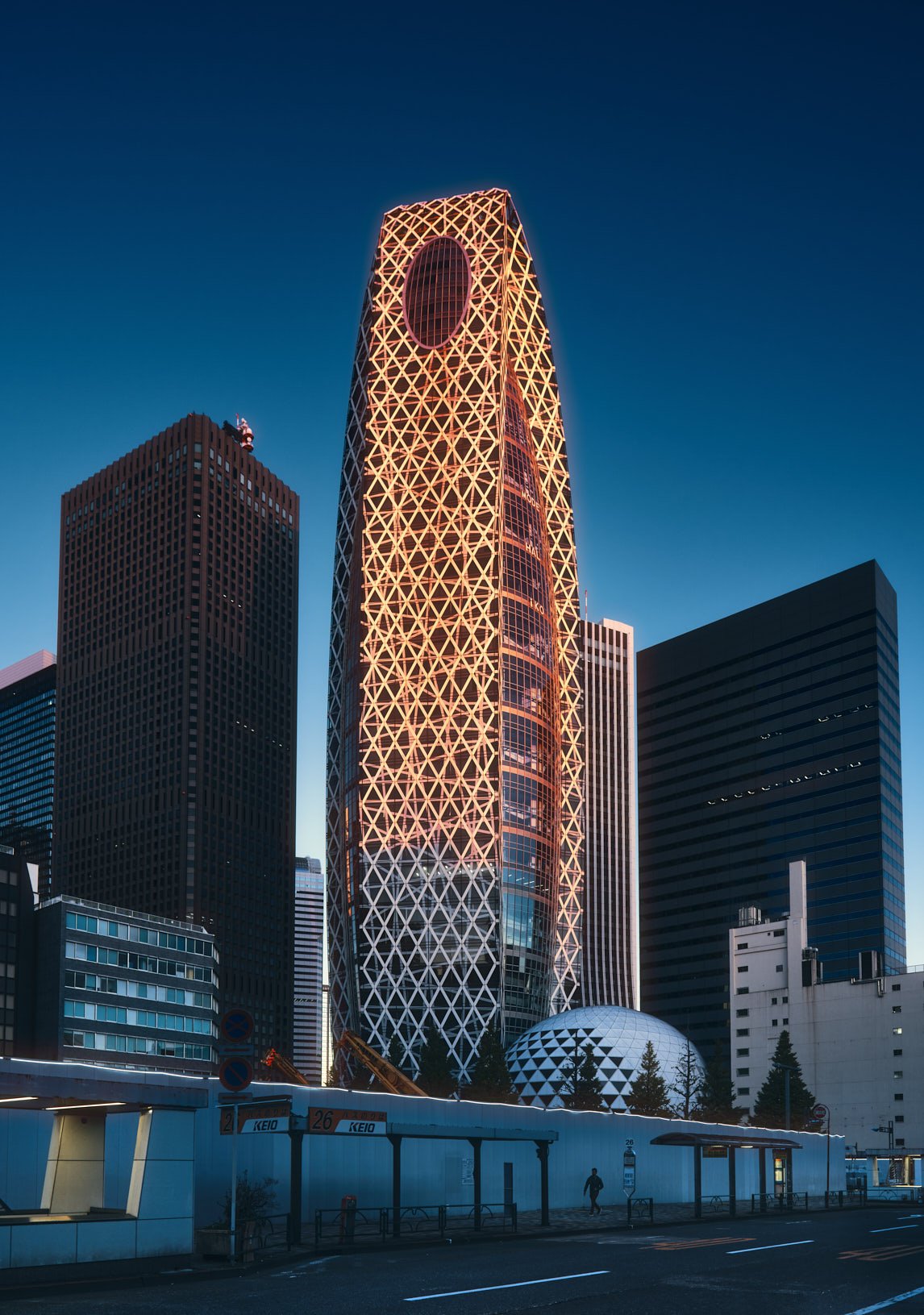 An architectural photograph of the Mode Cocoon Tower in Tokyo's Shinjuku district as morning sunlight kisses it's facade. A lone figure walks in shadow at it's base providing a striking sense of scale. Shot by Miami & Los Angeles based architectural