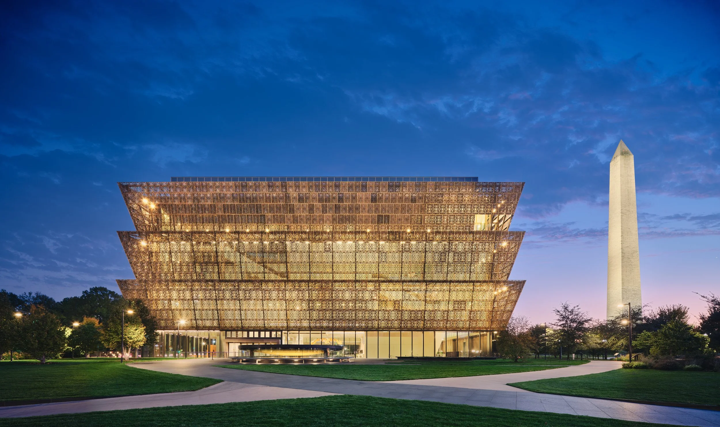 Architectural photograph of the Smithsonian National Museum of African American History and Culture in Washington, D.C., taken at twilight. The museum’s bronze lattice façade glows against a vibrant blue and purple sky, with the illuminated Washingto