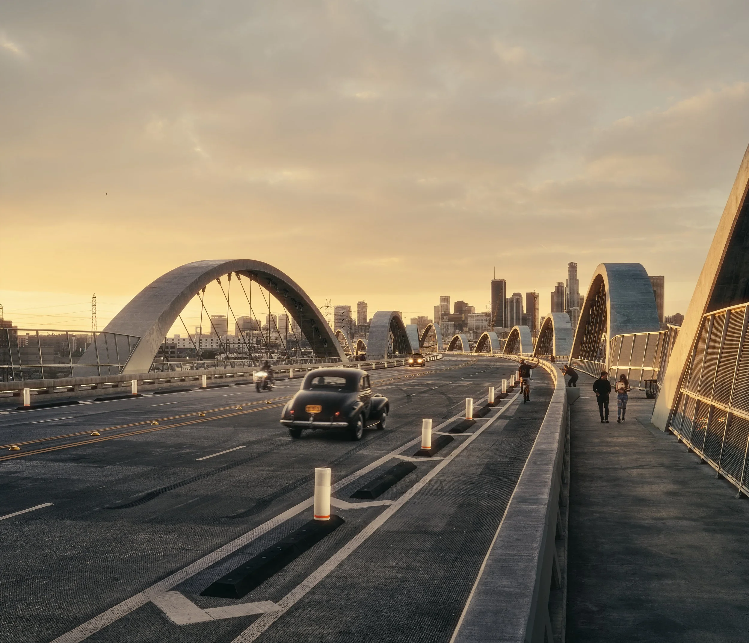 Architectural photograph of the 6th Street Viaduct in Los Angeles at sunset, featuring its signature arched supports stretching into the skyline. A classic car drives across the bridge while pedestrians and cyclists move along the adjacent path, capt