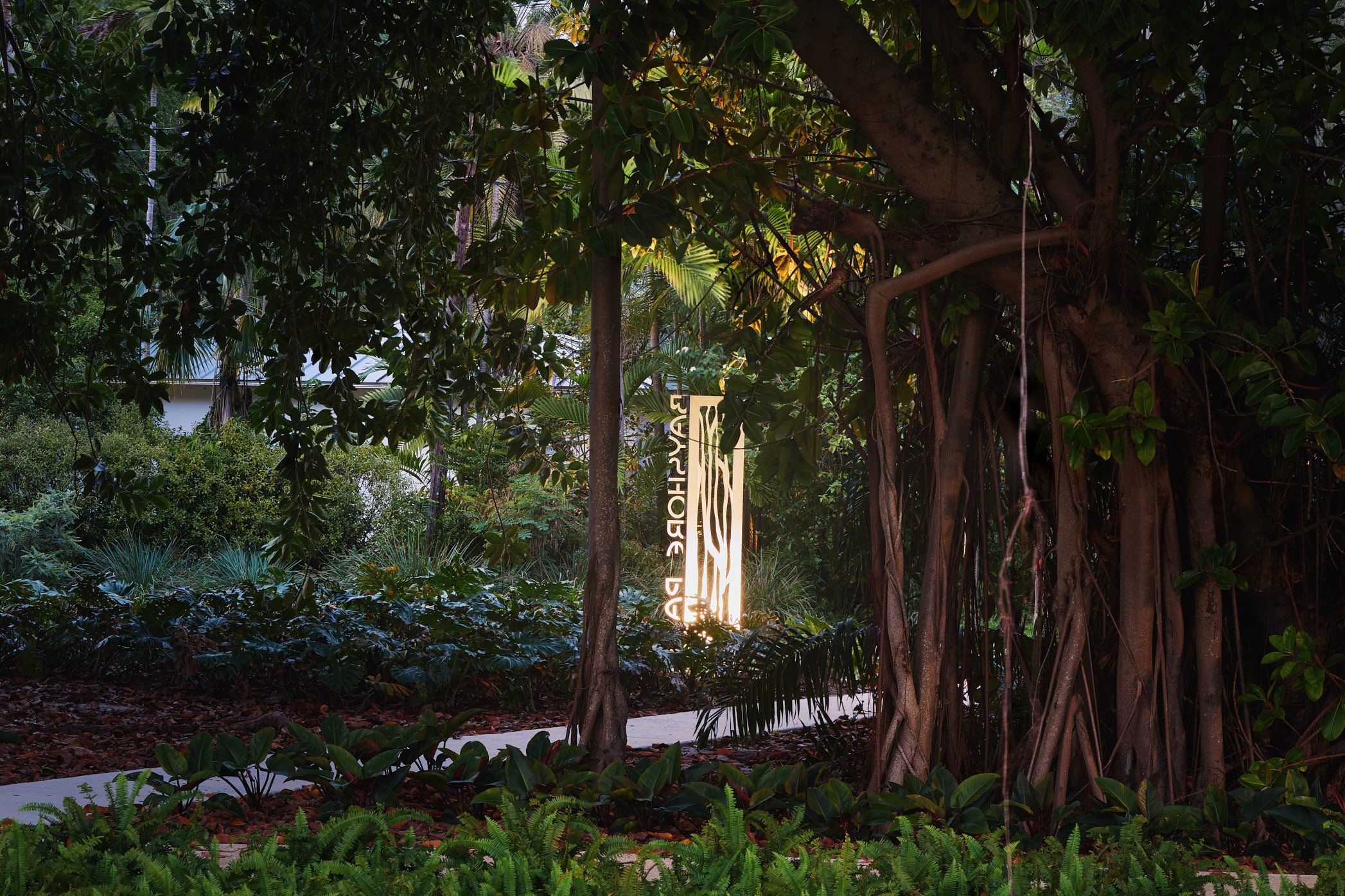 Architectural photograph of the illuminated Bayshore Park entrance sign framed by banyan trees and tropical plantings in Miami Beach, highlighting the landscape architecture and shaded garden pathways within the urban park. Captured by Miami and Los 