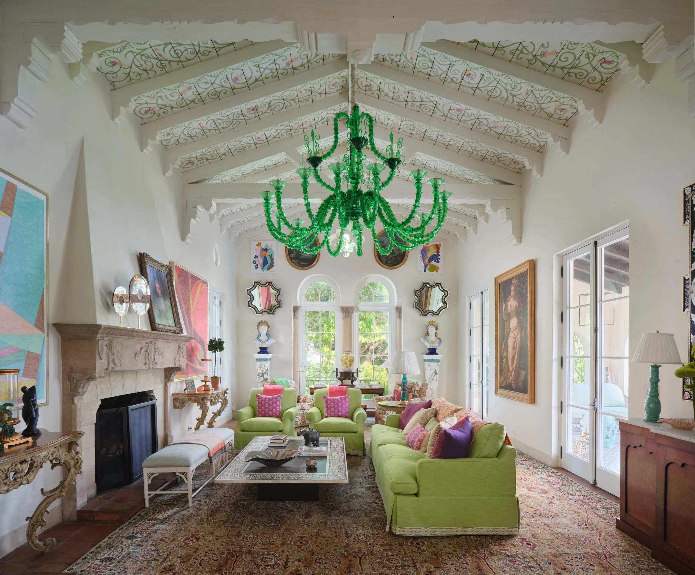 Architectural interiors photograph of Villa Serenita’s grand living room featuring a vaulted open-beam ceiling with painted floral panels, a striking emerald-green glass chandelier, an ornate carved stone fireplace, and bright lime green seating acce