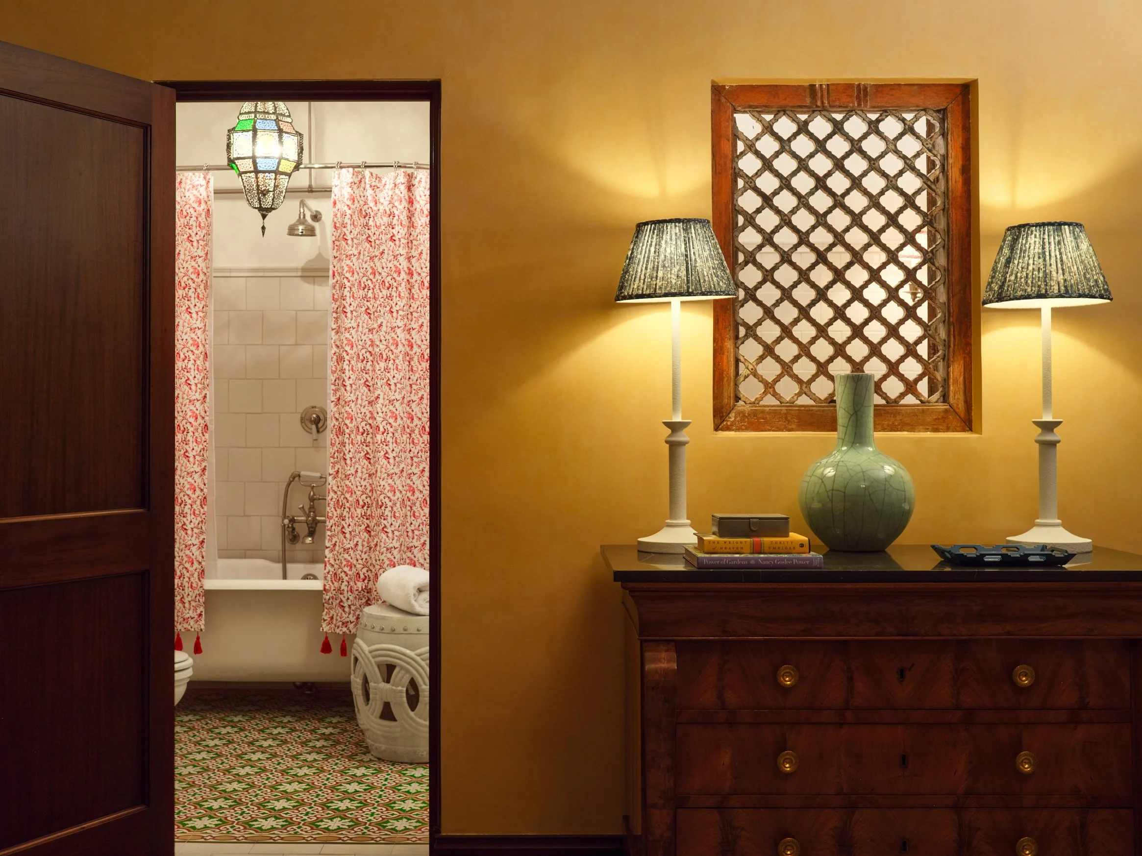 Architectural photograph of an elegant interior showing a warm yellow hallway with a wooden dresser, twin table lamps, and a ceramic vase, beneath a small, square Moroccan-inspired lattice window. To the left a wooden door opens into a bathroom with