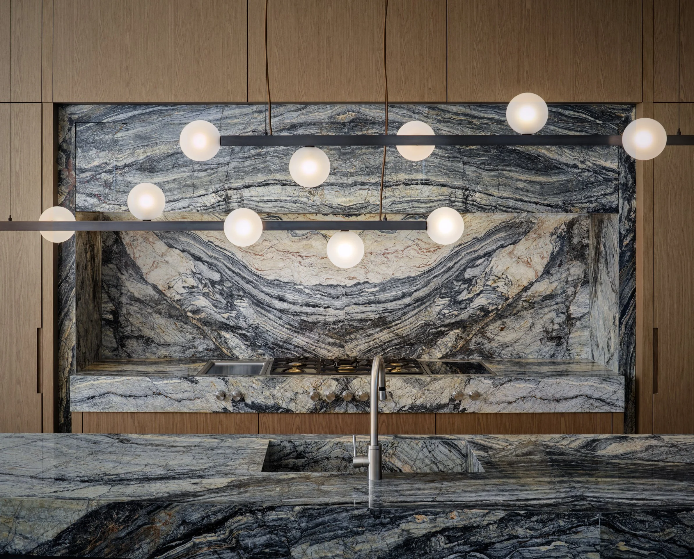 Architectural photograph of a luxury kitchen featuring dramatic bookmatched marble surfaces, integrated cooktop, and modern pendant globe lighting in a contemporary Miami residence. Captured by Miami and Los Angeles-based architectural and travel pho
