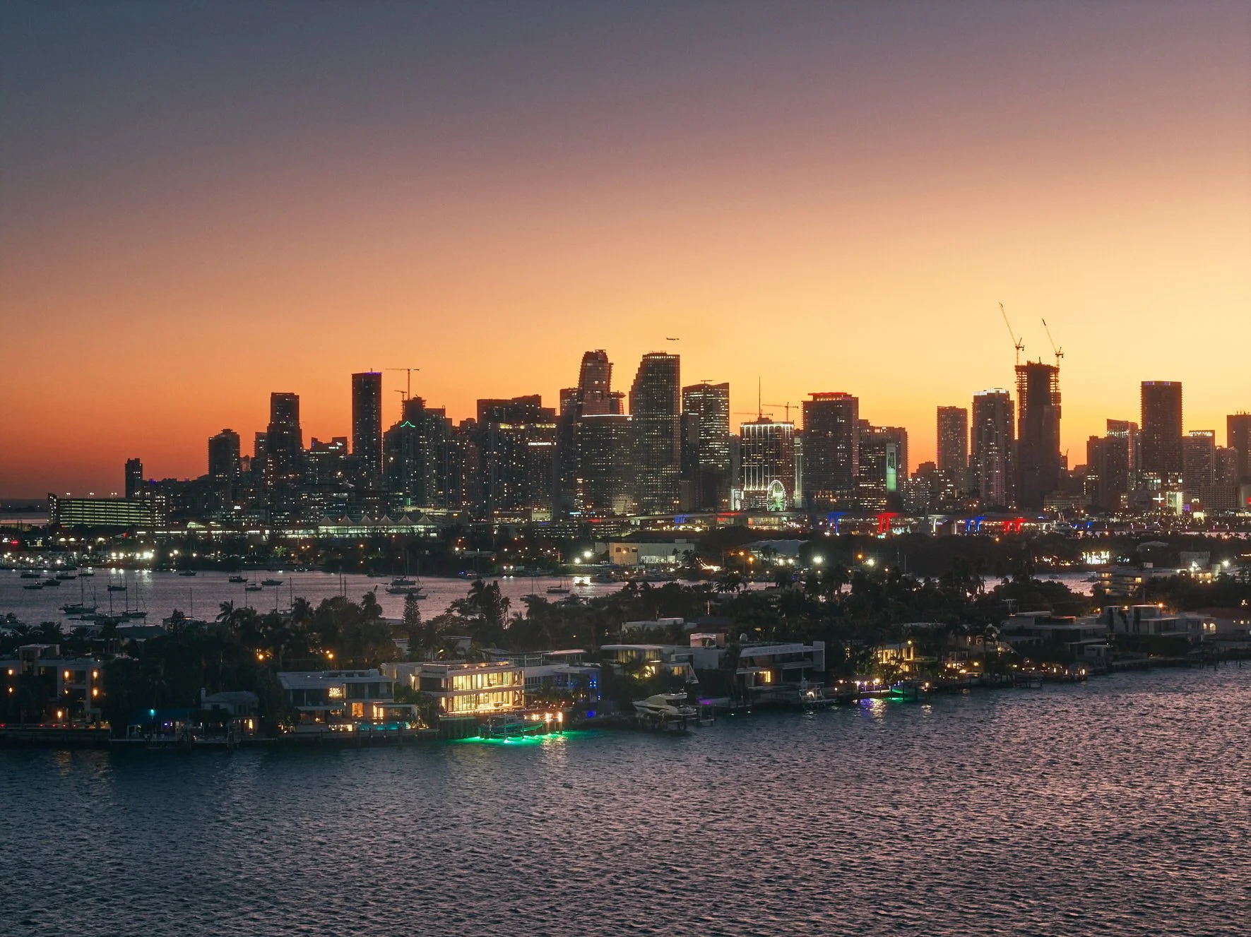 Architectural photograph of the Miami skyline at sunset viewed across Biscayne Bay from the Venetian Islands waterfront.  A lone waterfront residence stands illuminated amongst the rows of luxury homes. Captured by Miami and Los Angeles-based archite