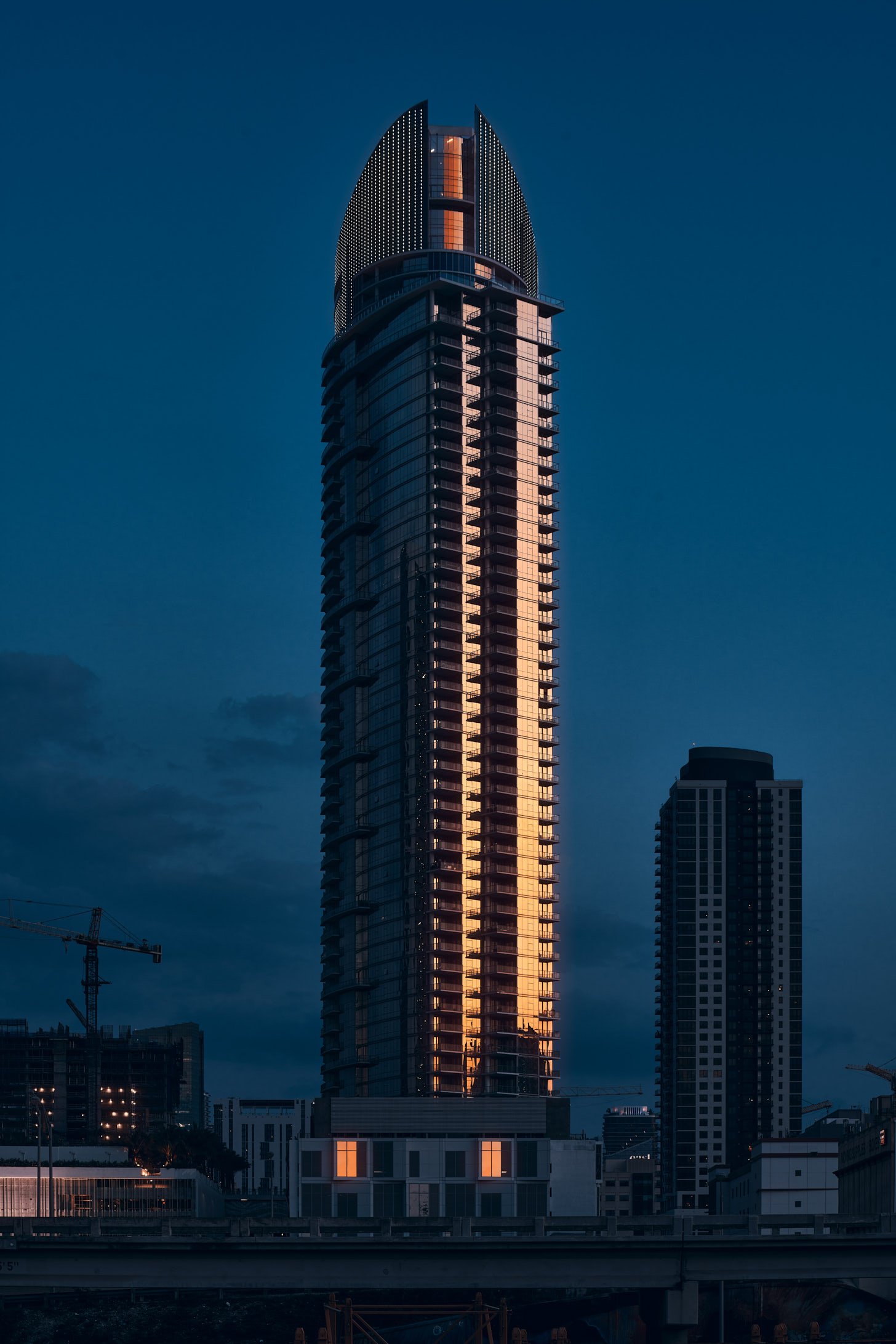 Architectural photograph of Paramount Miami tower at twilight, featuring a warm vertical reflection running up the building’s glass façade. The high-rise stands out against a dusky blue sky, with surrounding city structures and cranes in silhouette.