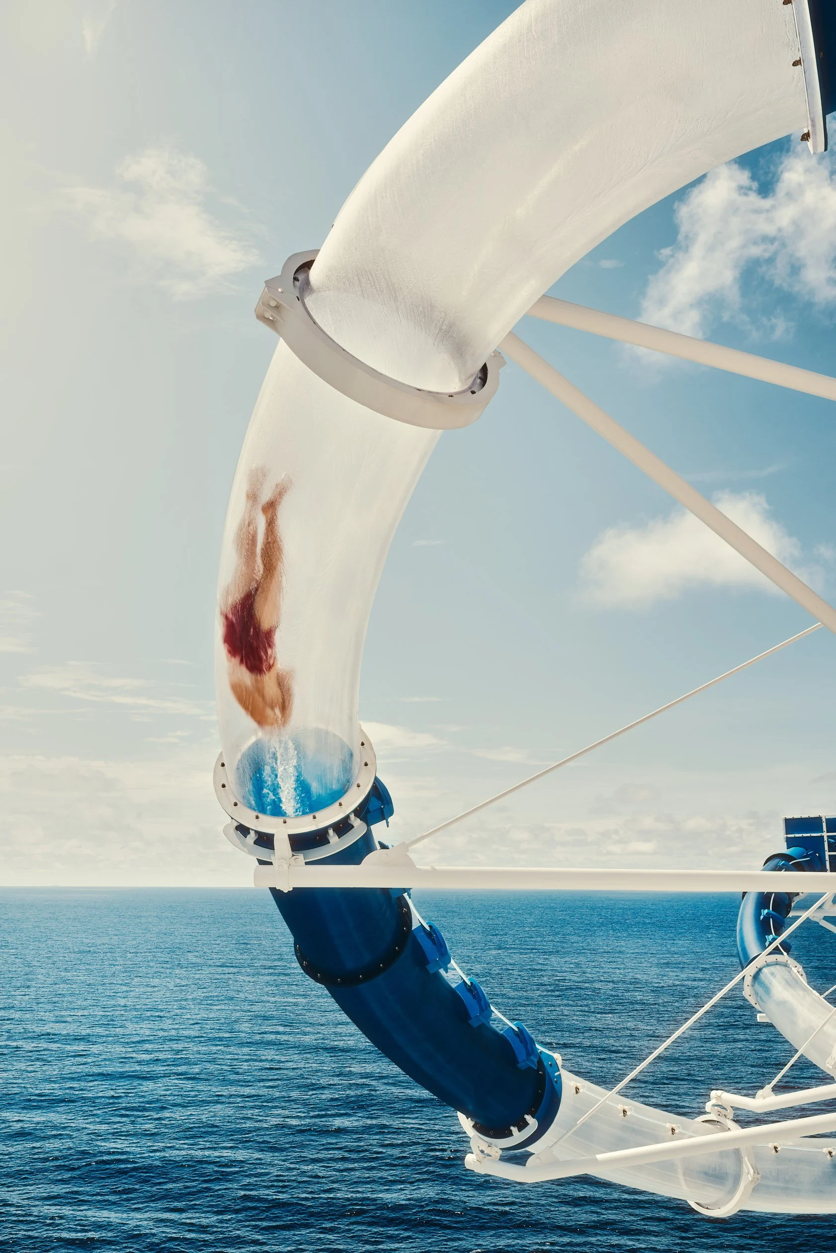 Architectural photograph of a guest mid-descent through the transparent waterslide extending over the open ocean aboard the Norwegian Cruise Line Bliss, highlighting the extraordinary structural engineering and experiential design of one of the world