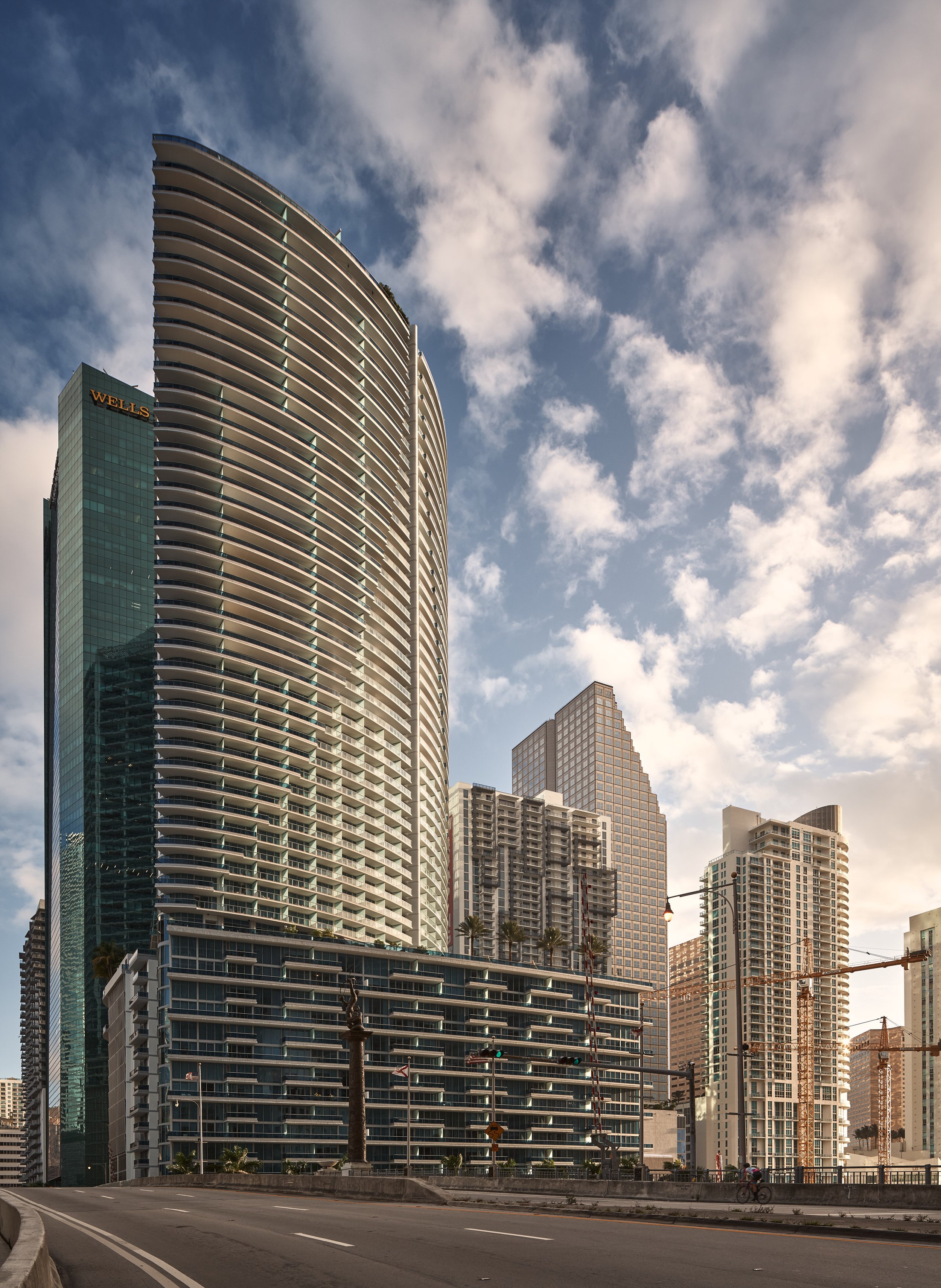 Architectural photograph of Epic Residences & Hotel in Brickell, Miami, with its distinctive curved facade catching soft morning light against a cloud-dappled sky. The image captures the skyline’s vertical rhythm, glass geometry, and the surrounding