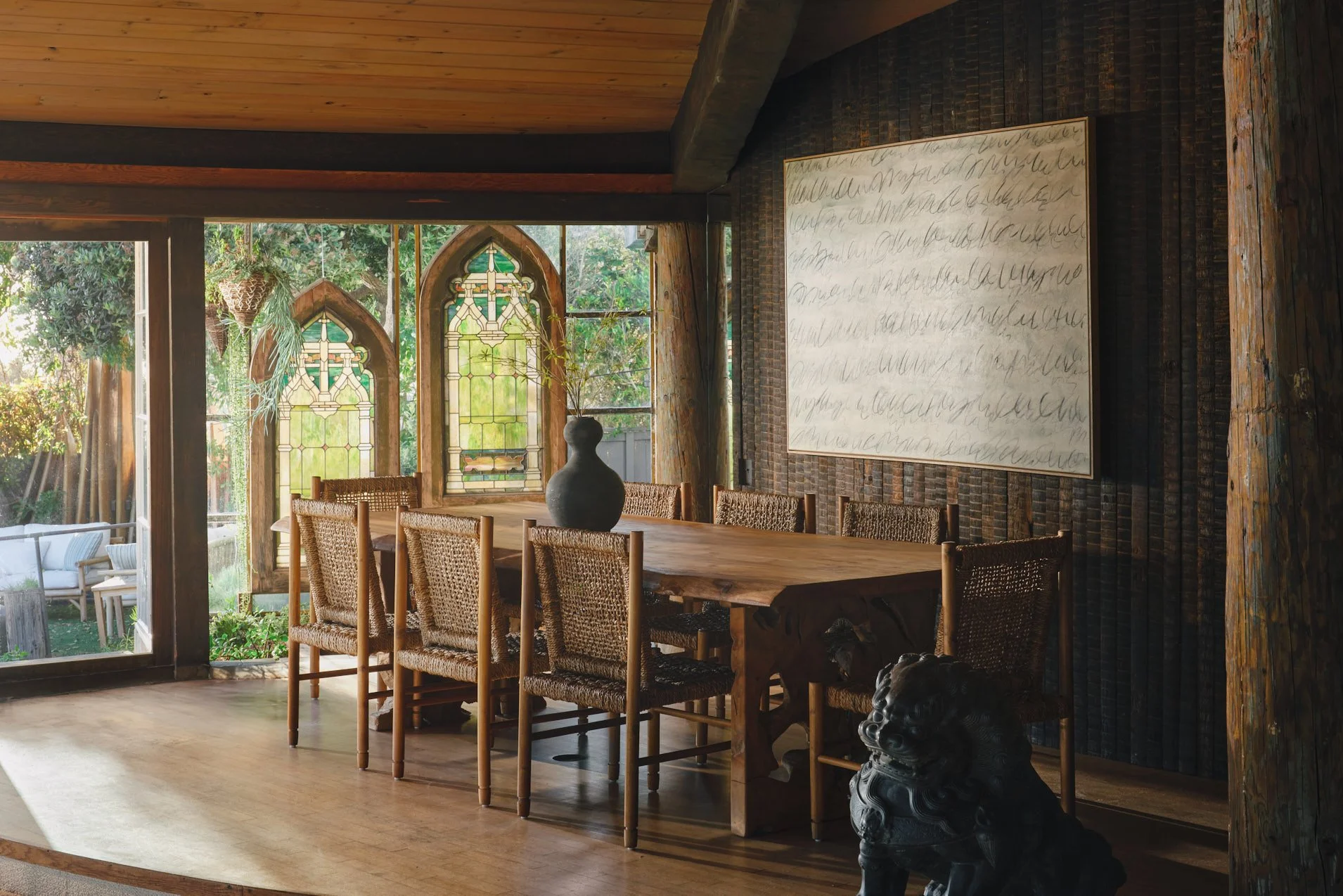 Architectural photograph of the Sandcastle House dining room in Malibu, featuring a live-edge wood table, woven chairs, and gothic stained glass windows repurposed as ocean-facing design elements. The space showcases Harry Gesner’s signature use of r