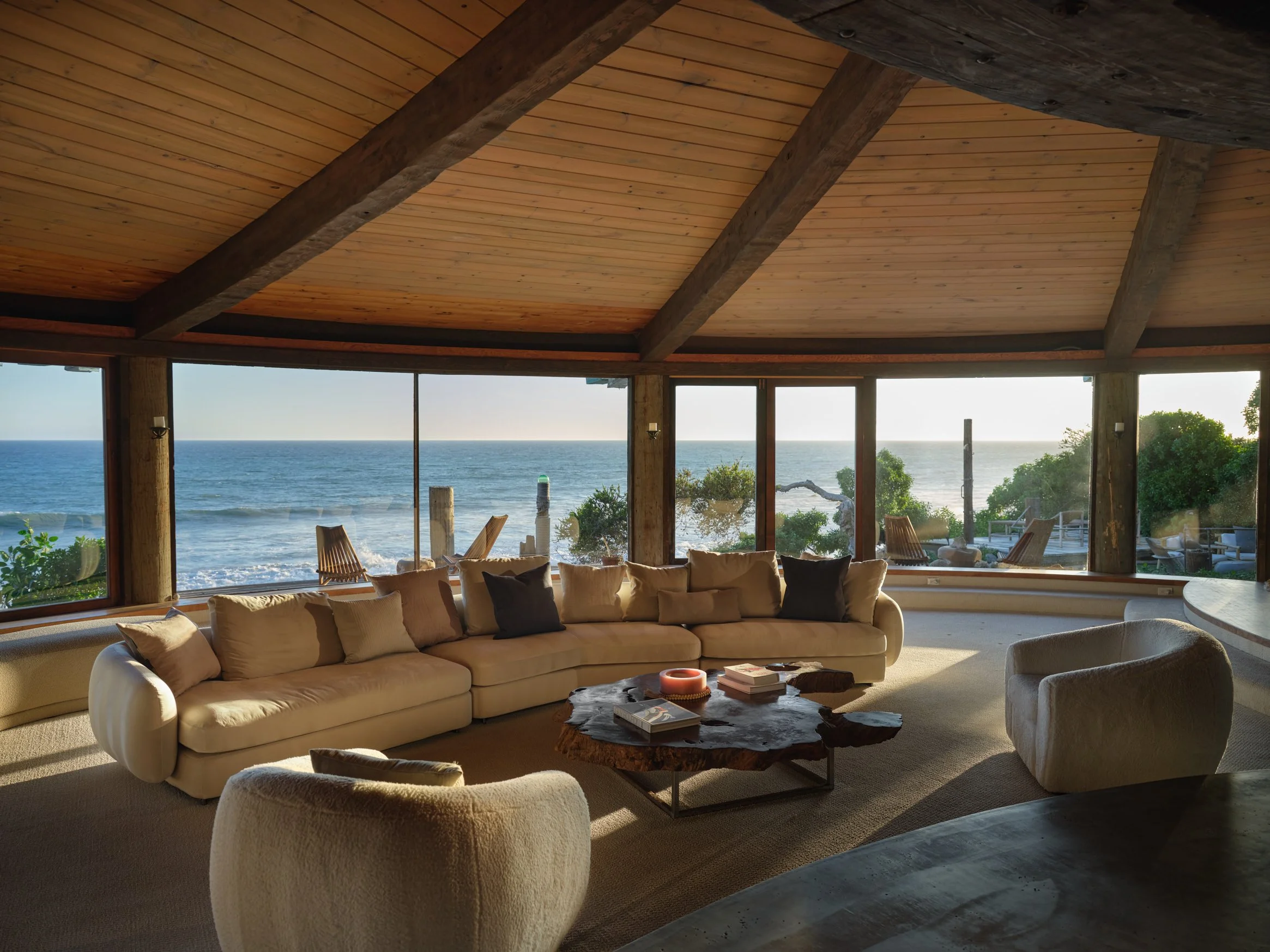 Architectural photograph of the Sandcastle House’s circular living room, showcasing panoramic ocean views through floor-to-ceiling glass. Exposed wooden beams radiate from the center, framing the curved cedar ceiling. Mid-century furnishings and a sc