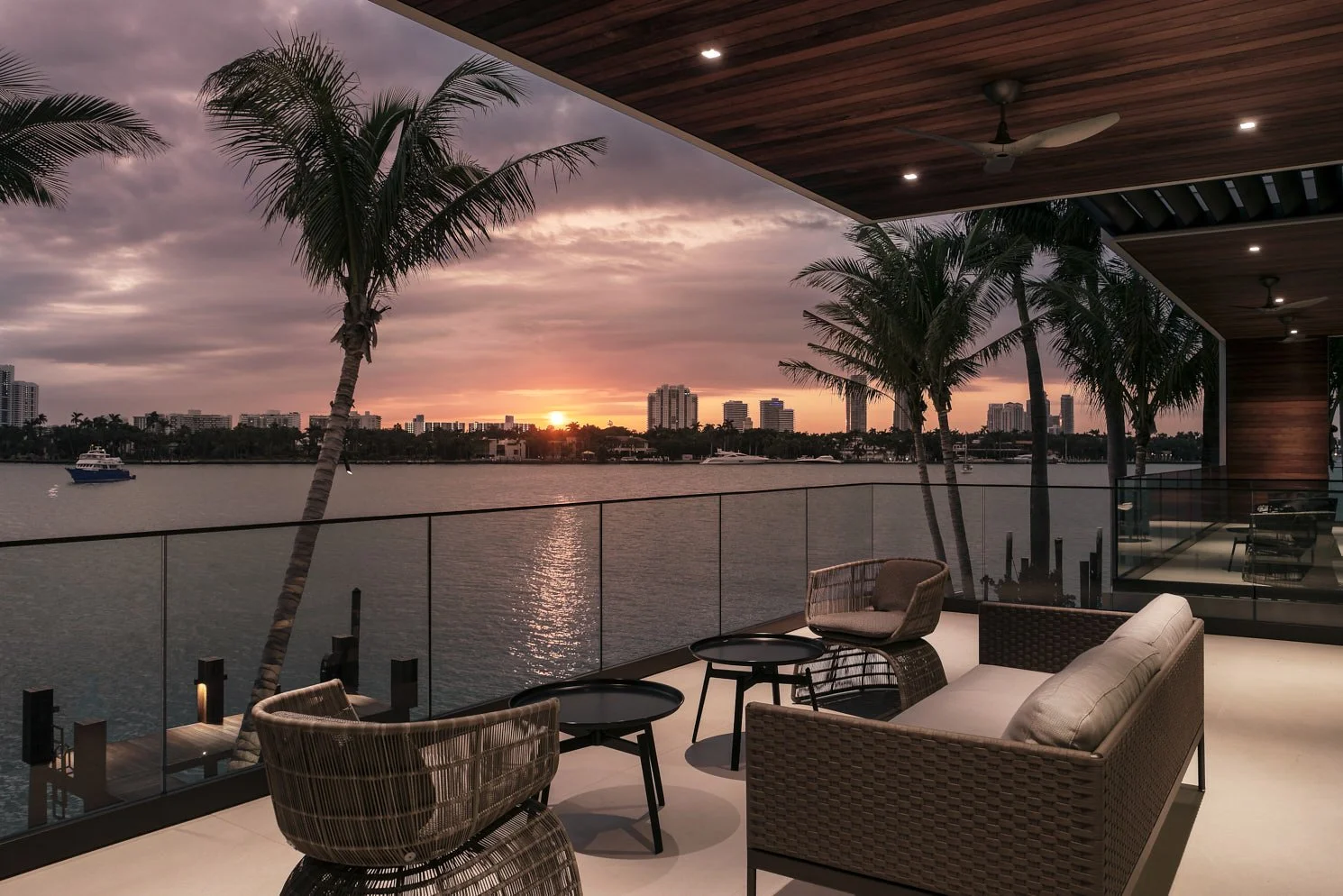 Architectural photograph of a luxury balcony overlooking Biscayne Bay at sunset on Star Island in Miami Beach, featuring modern wicker furniture, glass railings, and palm trees framing the waterfront skyline. Captured by Miami and Los Angeles-based a