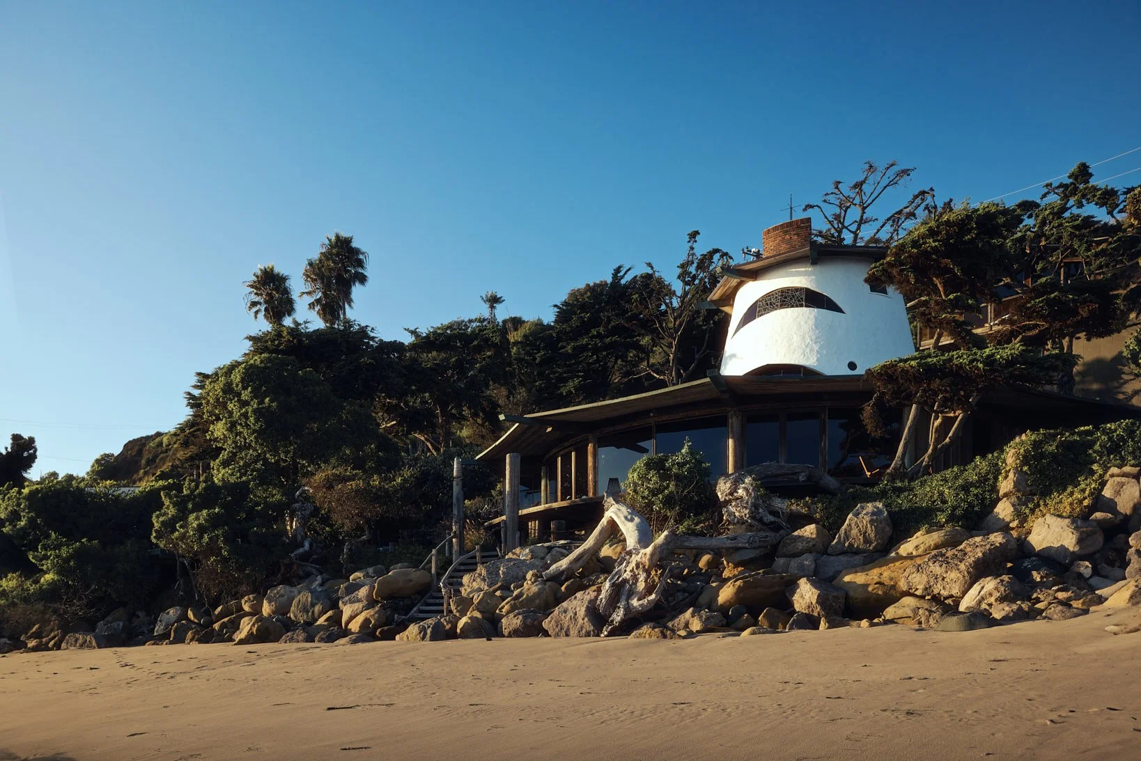 Architectural photograph of the iconic Sandcastle House in Malibu, designed by architect Harry Gesner. Shot from the beach at golden hour, the home’s sculptural tower rises above the oceanfront structure, flanked by native landscaping and dramatic ro