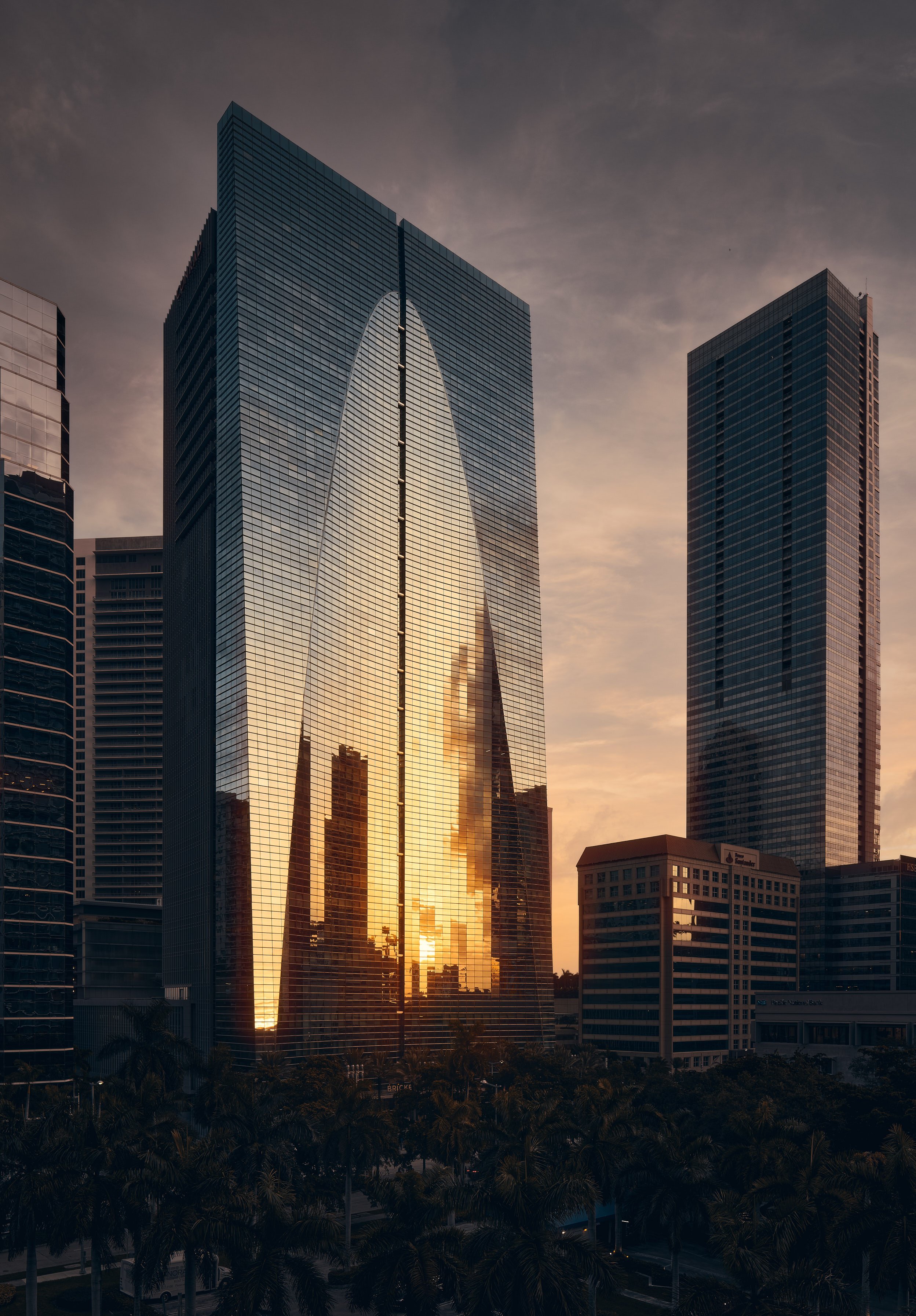 Architectural photograph of the Conrad Brickell building in Miami, Florida, featuring a stunning sunset reflection on its sleek glass façade. The dramatic silhouette of the Miami skyline is mirrored in the building’s surface, surrounded by other high