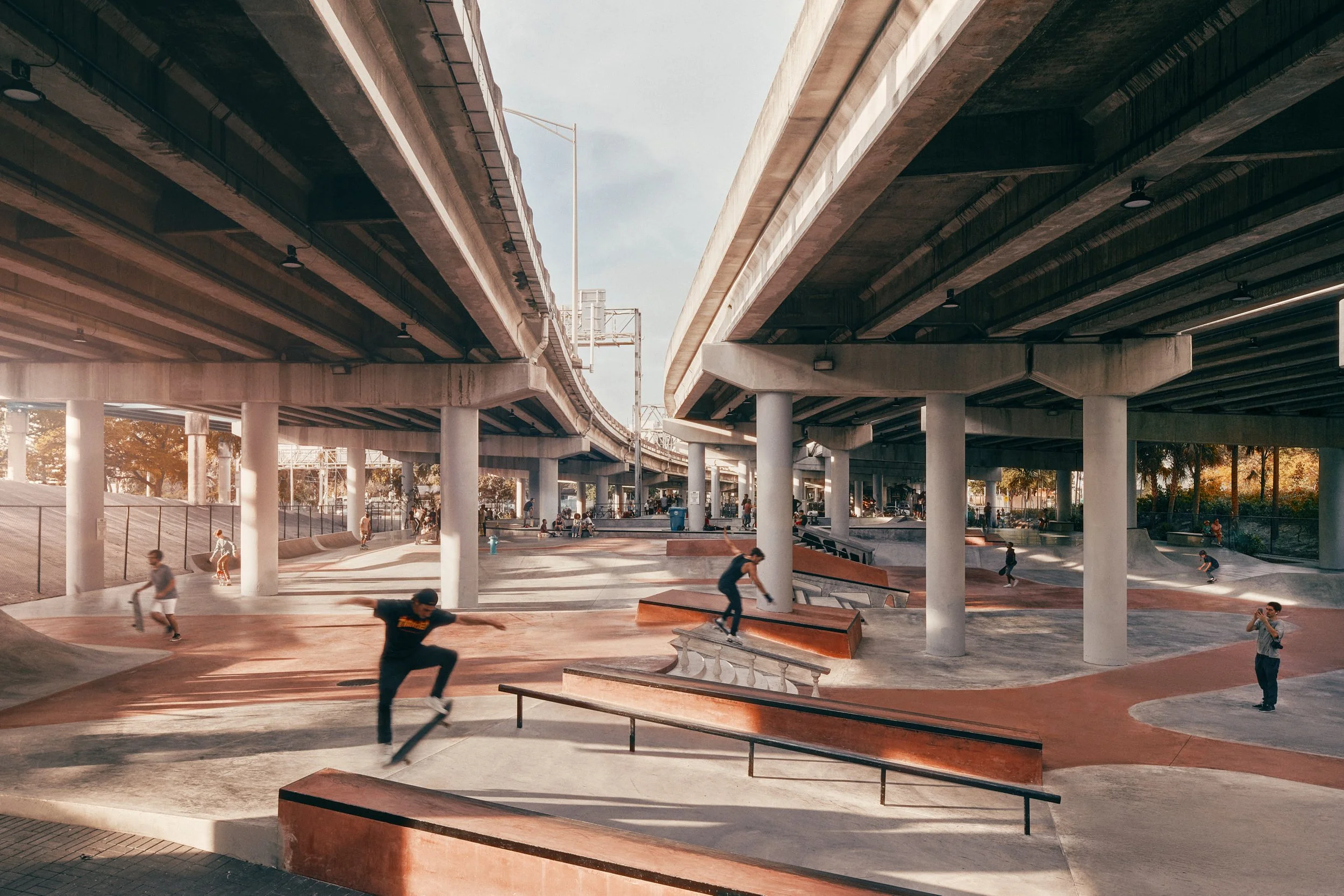 Architectural photograph of Lot 11 Skatepark in Miami, built beneath the soaring concrete infrastructure of the I-395 overpass, with skaters in motion across terracotta-toned ramps and rails under the dramatic colonnade of highway support columns — a