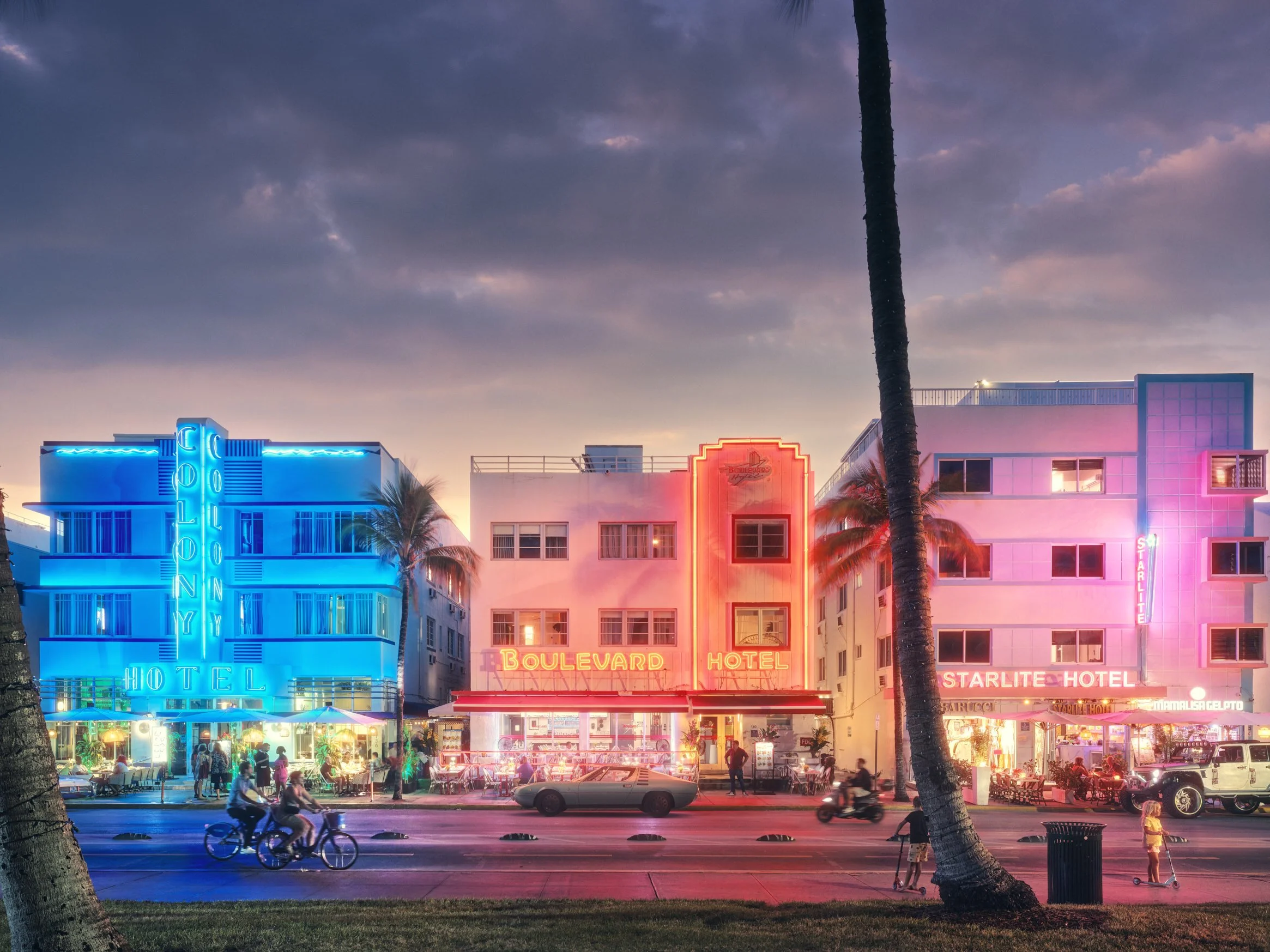 Architectural photograph of Ocean Drive’s iconic neon-lit hotels — the Colony Hotel, Boulevard Hotel, and Starlite Hotel — glowing at twilight in Miami’s South Beach. Palm trees and evening street activity add to the vibrant nightlife atmosphere of t