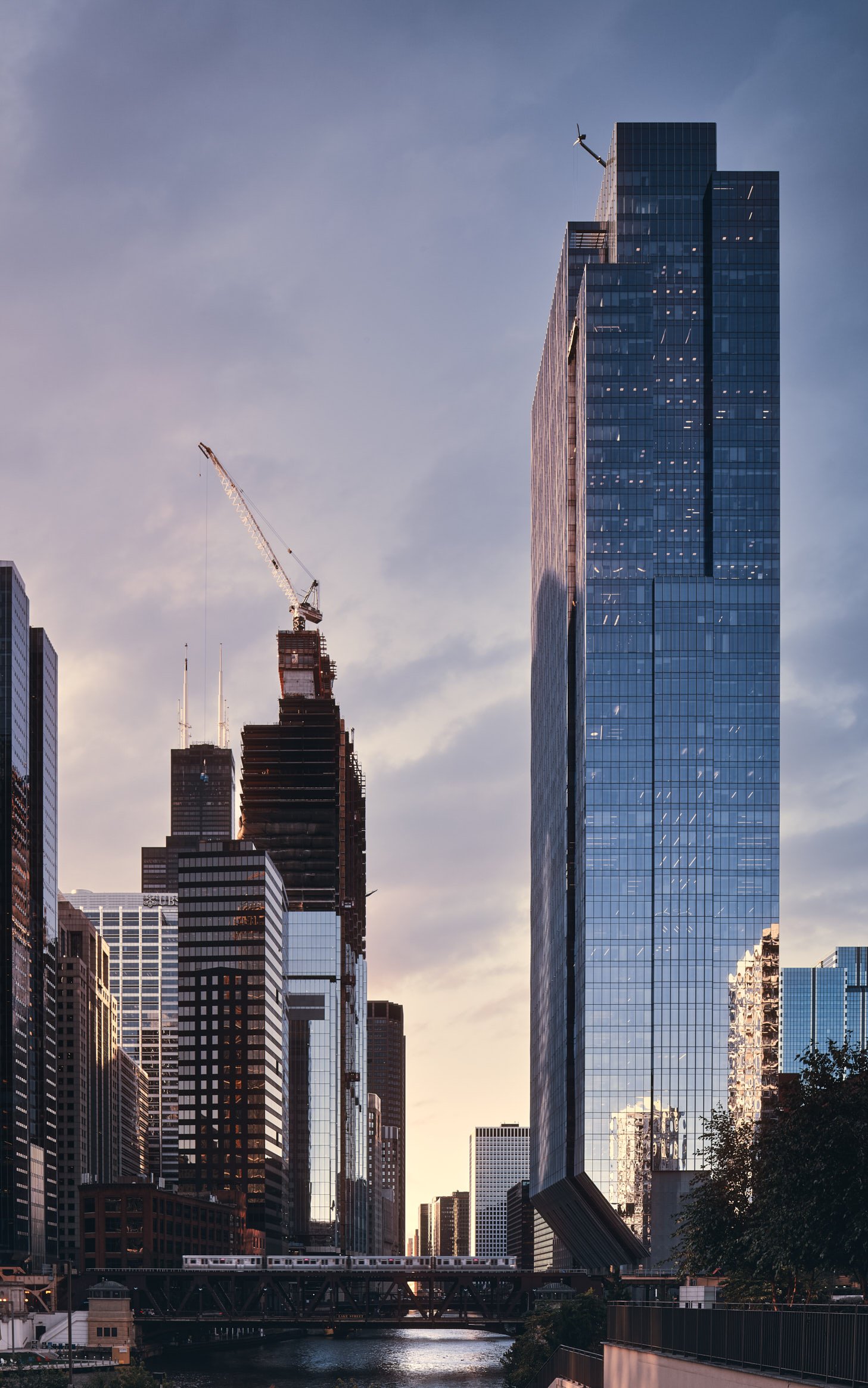 Architectural photograph of 150 North Riverside in Chicago, a striking cantilevered glass skyscraper set along the Chicago River, with an elevated train crossing in the foreground at sunset. The reflective facade mirrors the surrounding skyline under