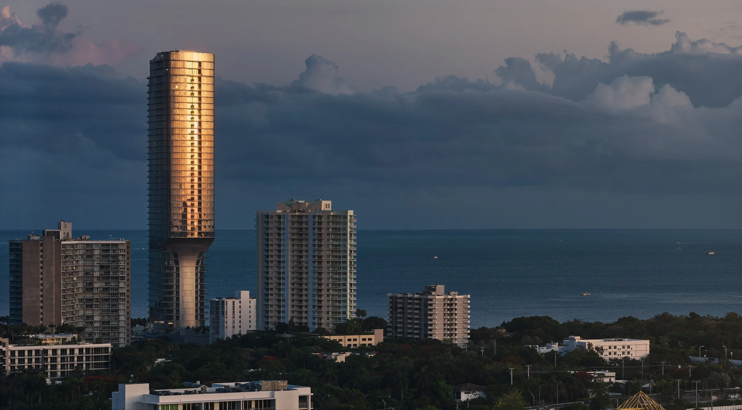 A dramatic aerial architectural photo of Una Residences in Miami's Brickell Neighborhood. Piercing sunset light paints the glass facade in a glowing orange ember as the blue waters of biscayne bay in the background harmonize the colors. Photographed 