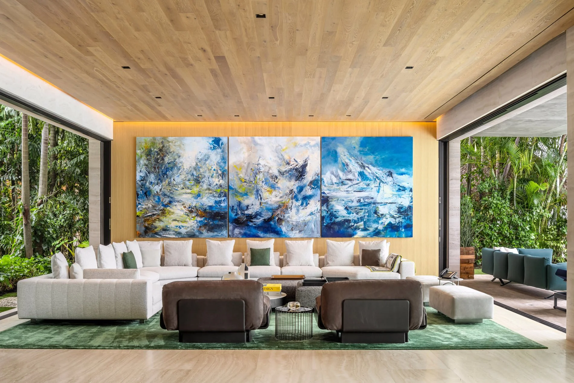Architectural photograph of a luxurious modern Miami living room with sliding glass walls opening to lush tropical greenery. The space features a long white sectional sofa accented by neutral pillows, two dark leather chairs, and a bold triptych of a