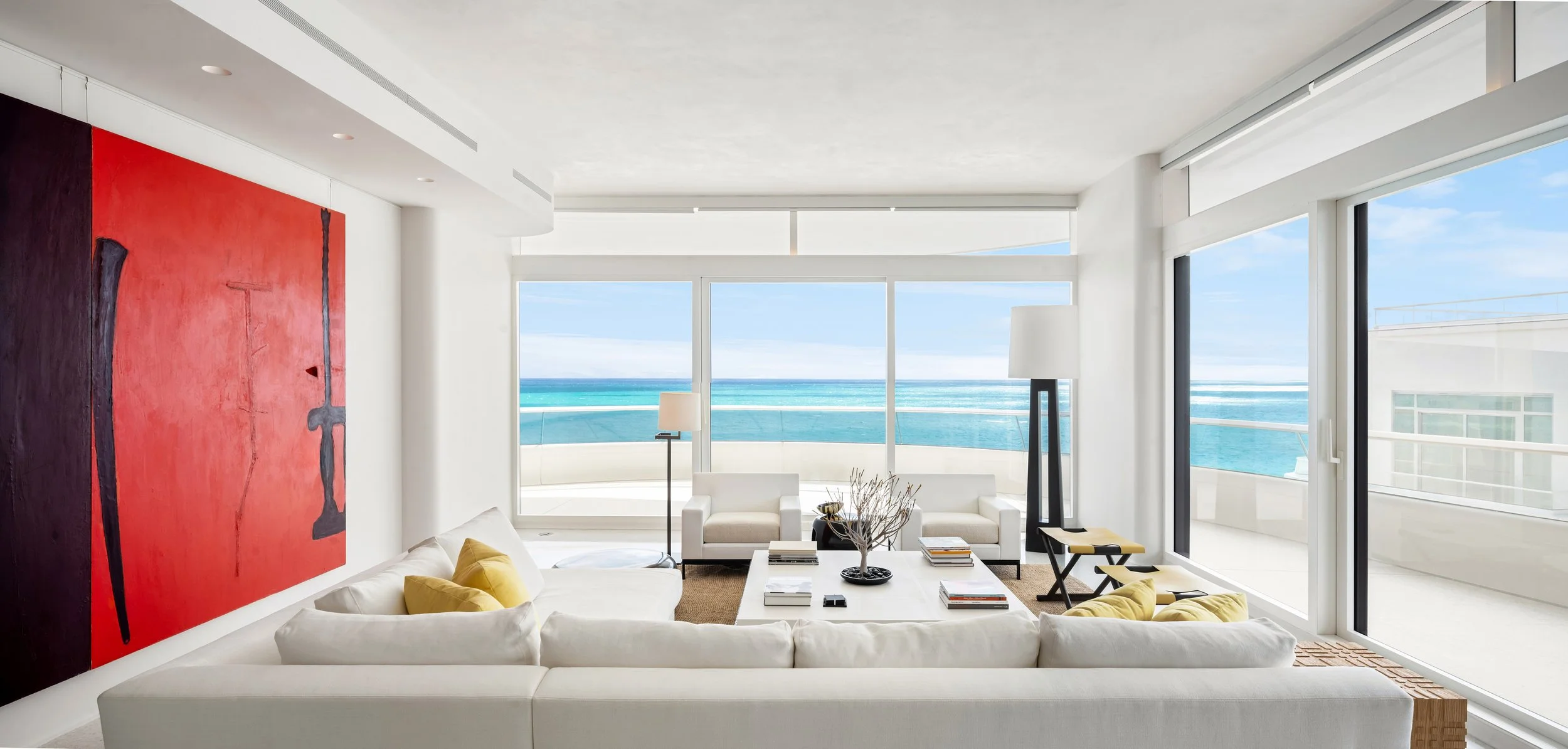 Architectural photograph of a modern oceanfront living room in Faena House, featuring floor-to-ceiling windows with sweeping views of the turquoise sea. The interior is bright and minimal with white walls, a bold red abstract painting, clean furnitur