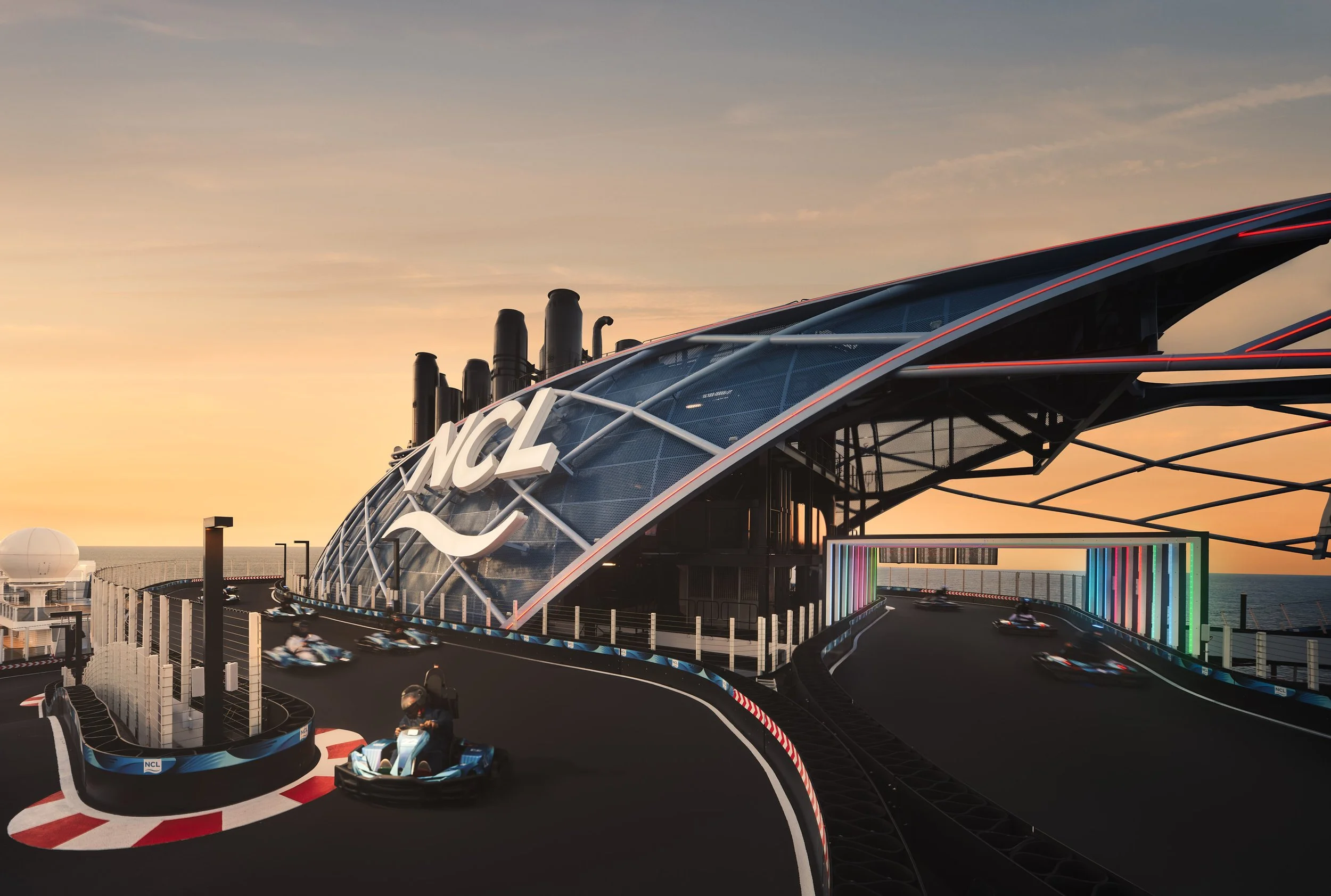 Architectural photograph of the go-kart racetrack aboard the Norwegian Cruise Line Prima at twilight, featuring the dramatic NCL-branded canopy structure, motion-blurred karts in action, and a vibrant color-lit gateway against an open ocean horizon —