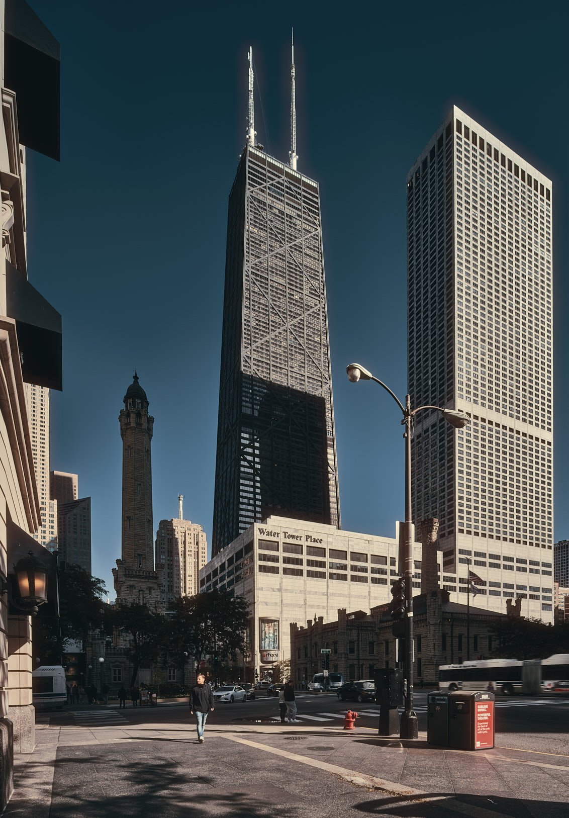 Architectural photograph of the John Hancock Center in Chicago, showcasing the iconic X-braced skyscraper rising behind Water Tower Place on a crisp, clear day. The historic Chicago Water Tower stands nearby, anchoring the image with a blend of moder