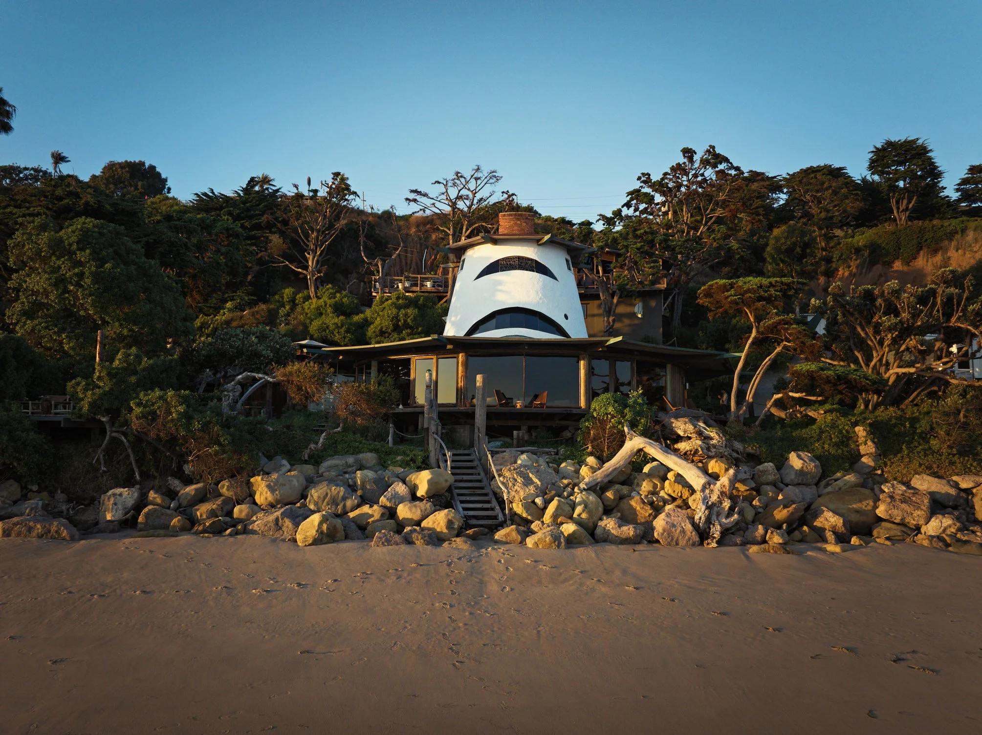 Architectural photograph of the iconic Sandcastle House in Malibu, designed by architect Harry Gesner. Shot from the beach at golden hour, the home’s sculptural tower rises above the oceanfront structure, flanked by native landscaping and dramatic ro