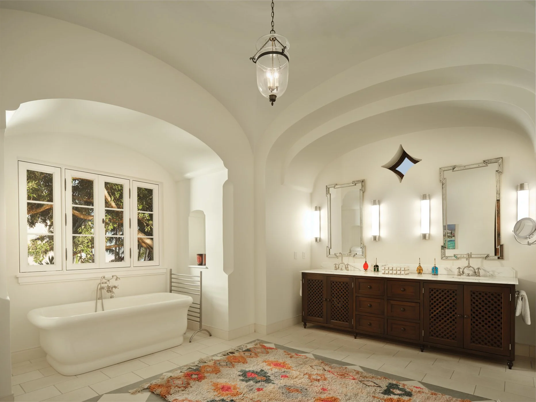 Architectural photograph of a spacious Mediterranean-style bathroom featuring a freestanding white soaking tub beneath a wide multi-pane casement window framed in white, with views of lush greenery outside. The room has a vaulted ceiling with soft cu