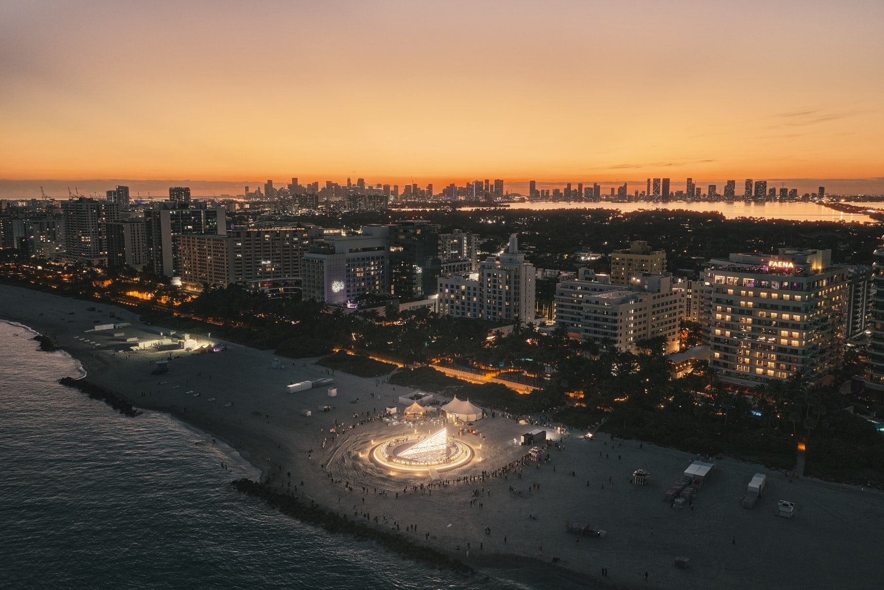 Architectural photograph of Es Devlin’s Library of Us installation at Faena Miami Beach during Art Basel Miami Beach, captured at dusk with the illuminated circular structure set against the Miami skyline. The aerial composition emphasizes the relati