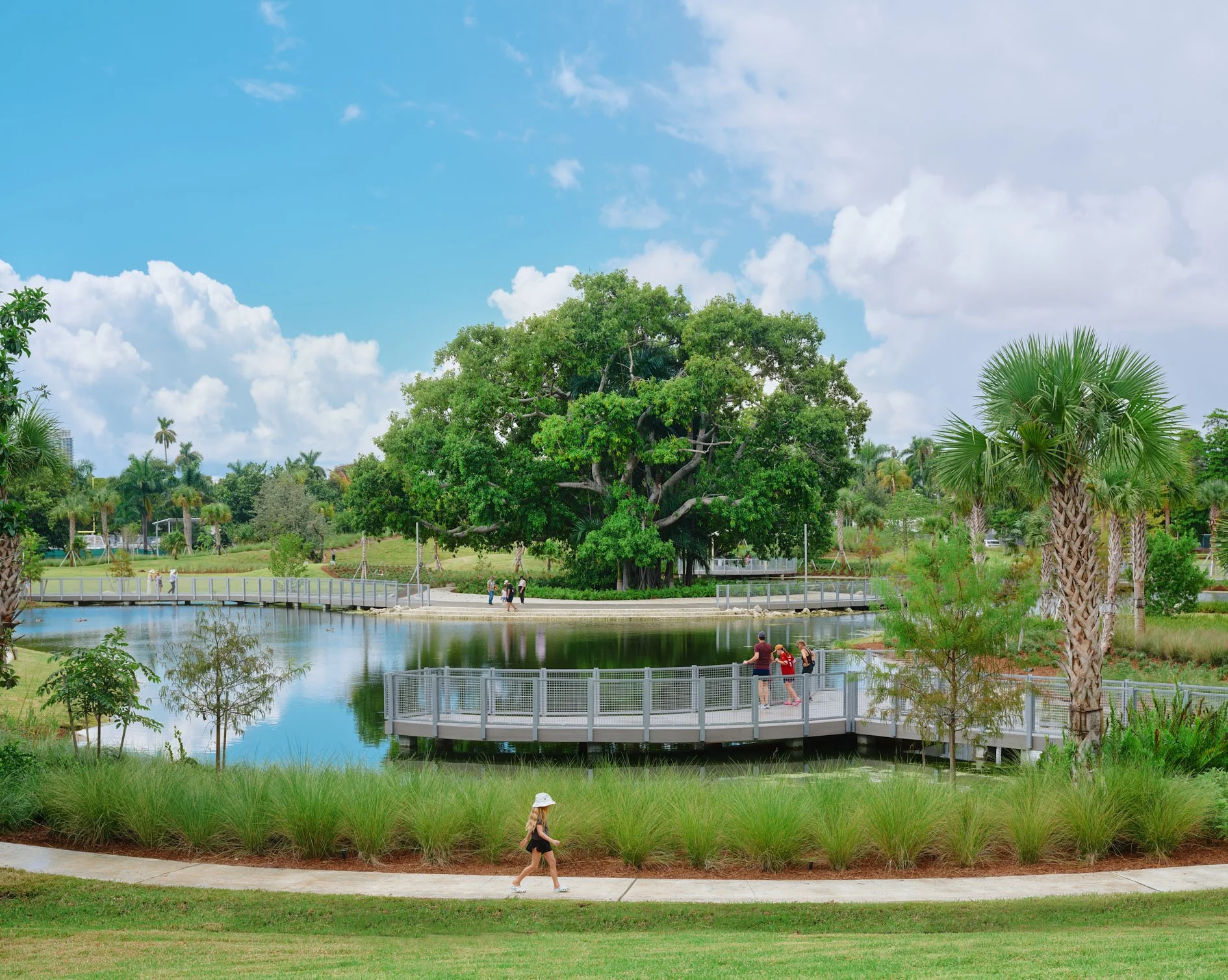 Architectural photograph of the banyan tree island at Bayshore Park in Miami Beach, Florida, surrounded by a reflective lake and connected by pedestrian boardwalks, with visitors enjoying the 19.4-acre civic park designed by Savino Miller — one of Mi