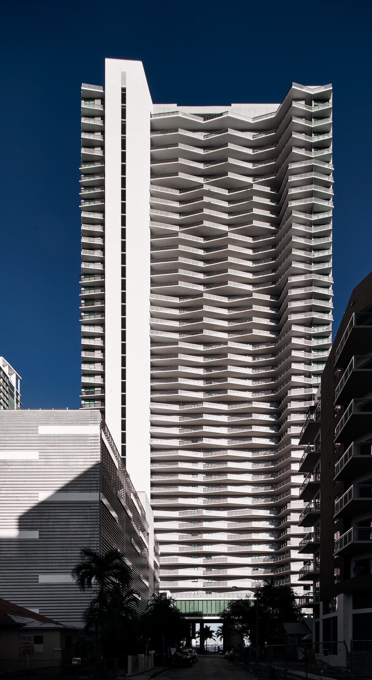 Architectural photograph of Icon Bay condominium in Miami, Florida, showcasing its distinctive zig-zag balcony design under crisp sunlight and deep blue sky. The high-rise structure is framed symmetrically between shadowed buildings, with palm trees
