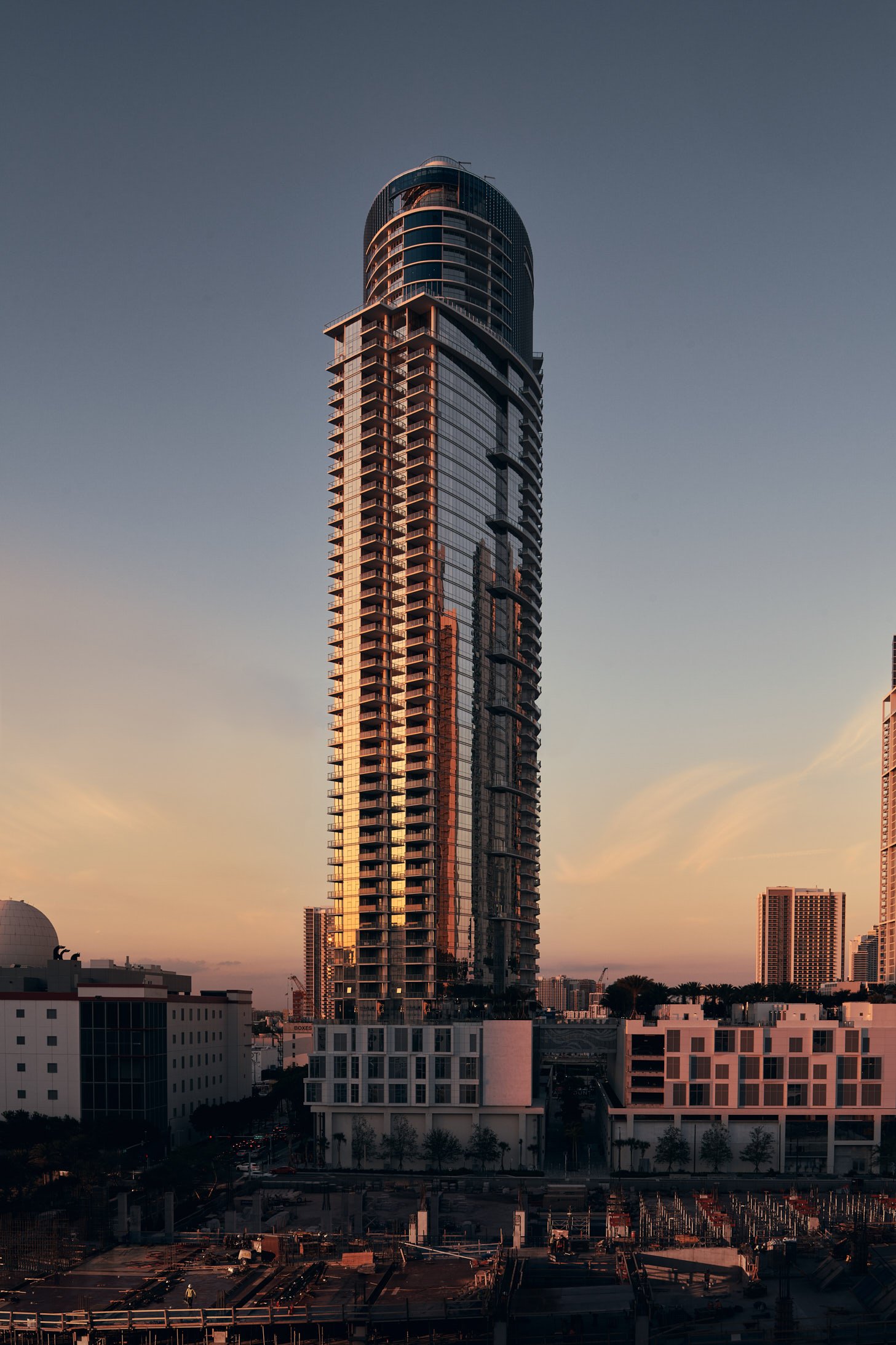 Architectural photograph of the Paramount Miami Worldcenter tower at sunset, its glass façade reflecting warm golden light against a pastel sky. The modern high-rise is surrounded by mid-rise buildings and construction, showcasing Miami’s evolving sk