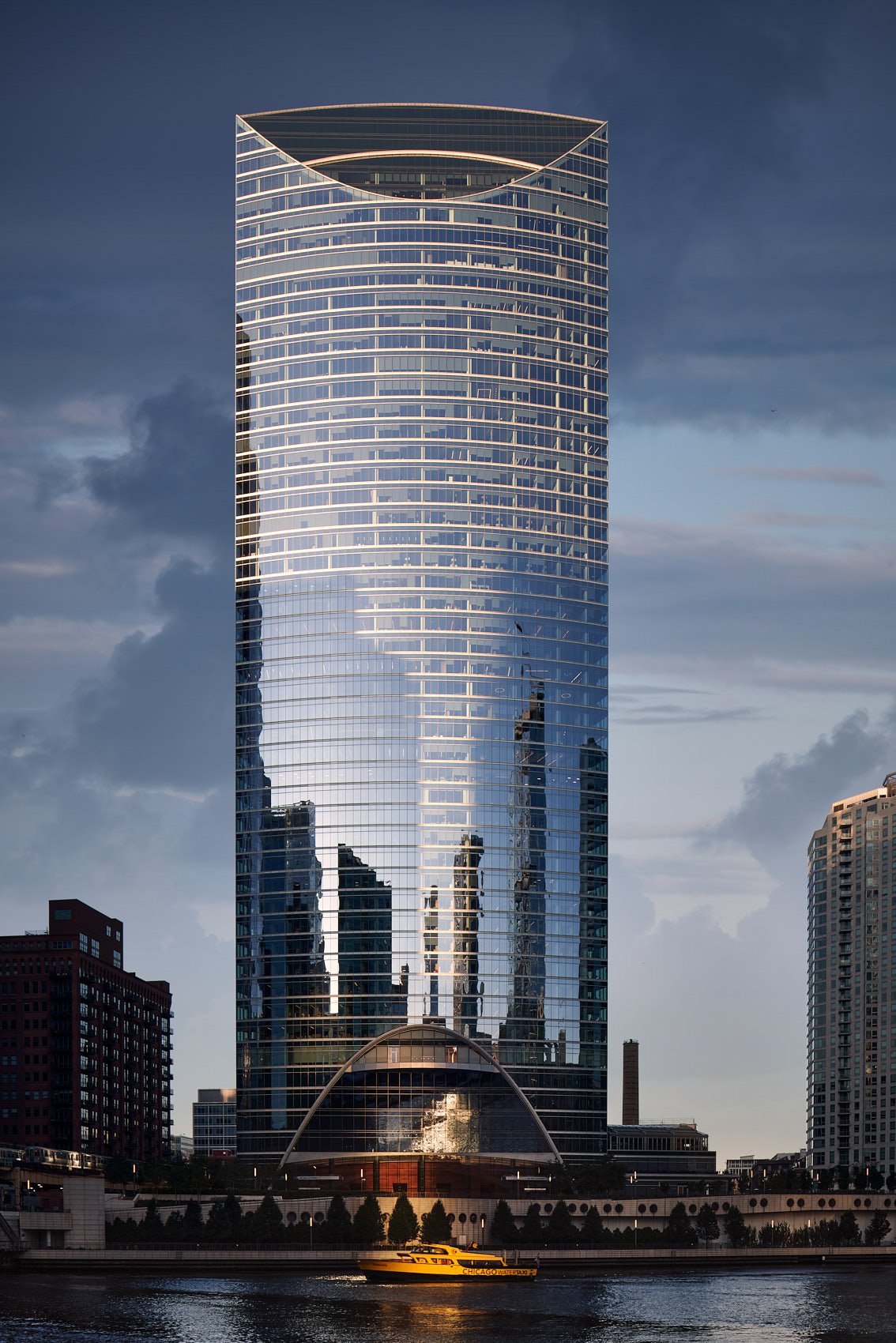 Architectural photograph of River Point, a modern high-rise in downtown Chicago with a curved glass façade reflecting the city skyline. A bright yellow water taxi floats on the Chicago River beneath the tower, with dramatic clouds and evening sunligh
