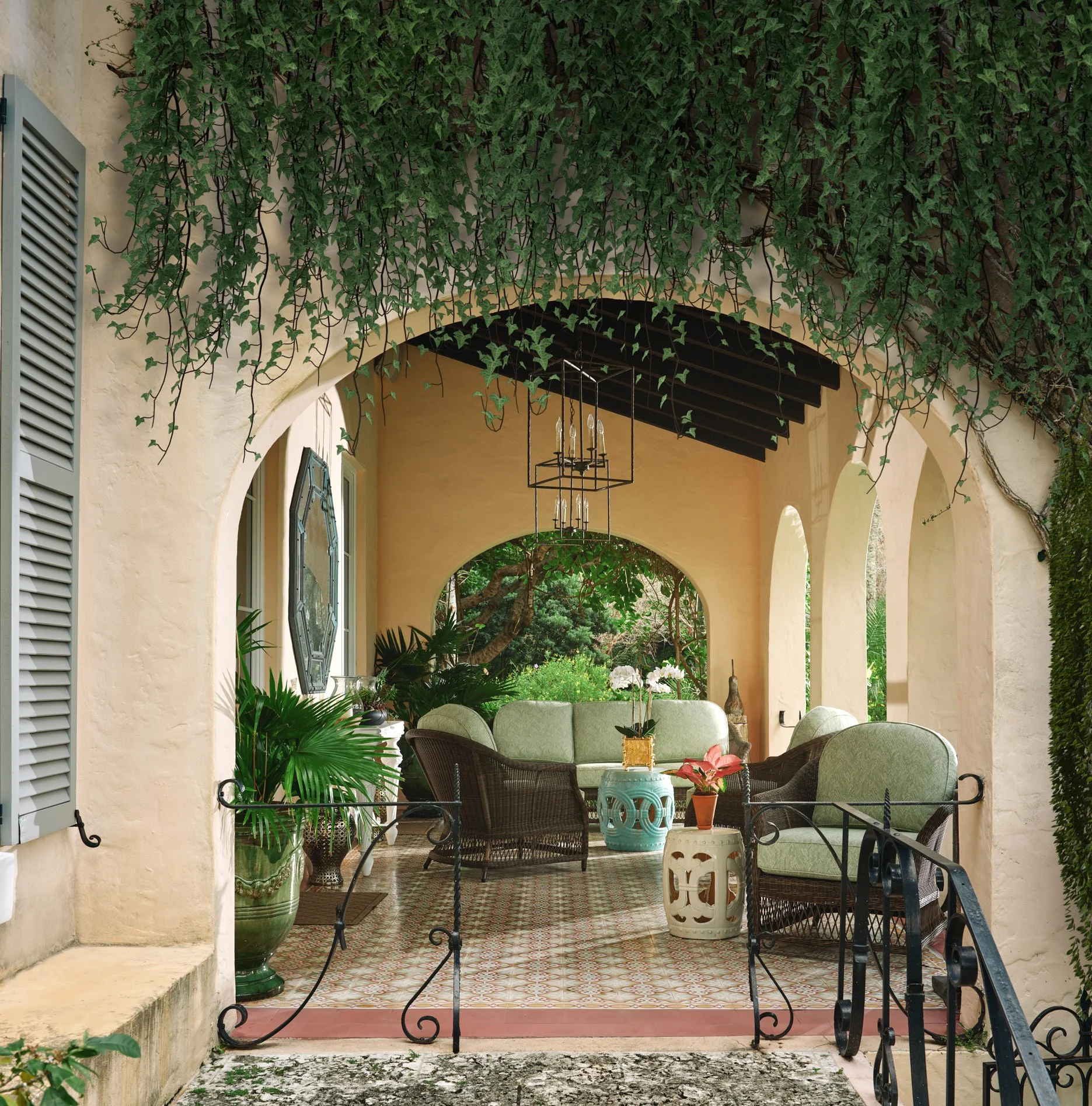 Architectural photograph of a Mediterranean-style covered terrace framed by soft peach stucco arches, with cascading ivy dripping from above. The spaces features patterned tile flooring, wrought iron railings, wicker seating with sage green cushions,