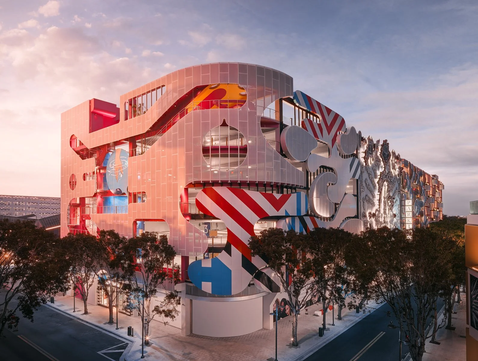 Architectural photograph of Museum Garage in the Miami Design District at twilight, showcasing the building's iconic multi-artist facade featuring bold graphic murals, sculptural lettering, circular cutouts, and vibrant color panels — one of Miami's 