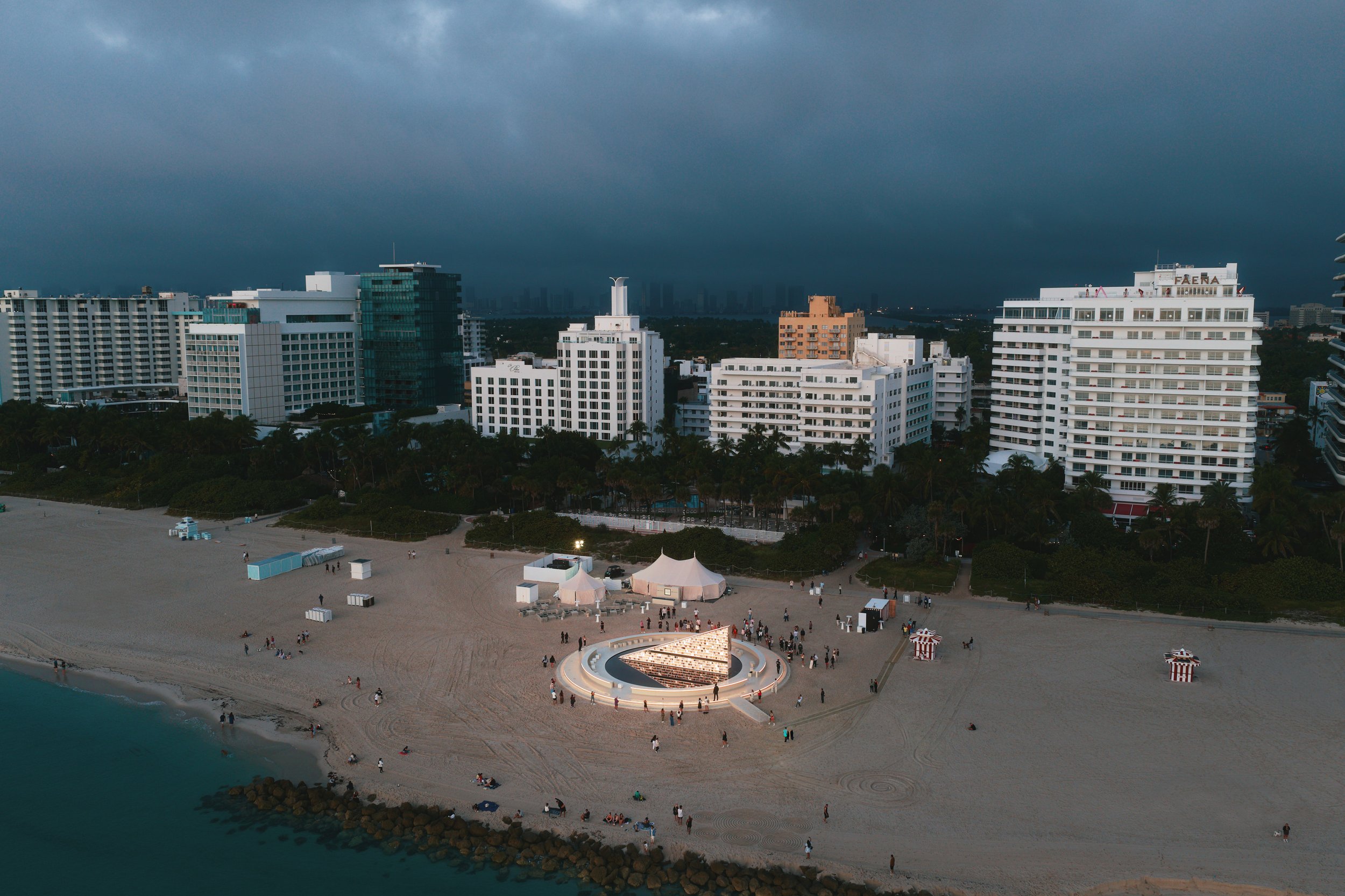 Architectural photograph of Es Devlin’s Library of Us public art installation at Faena Miami Beach during Art Basel Miami Beach, captured from an aerial perspective as the illuminated circular structure sits along the shoreline beneath approaching st