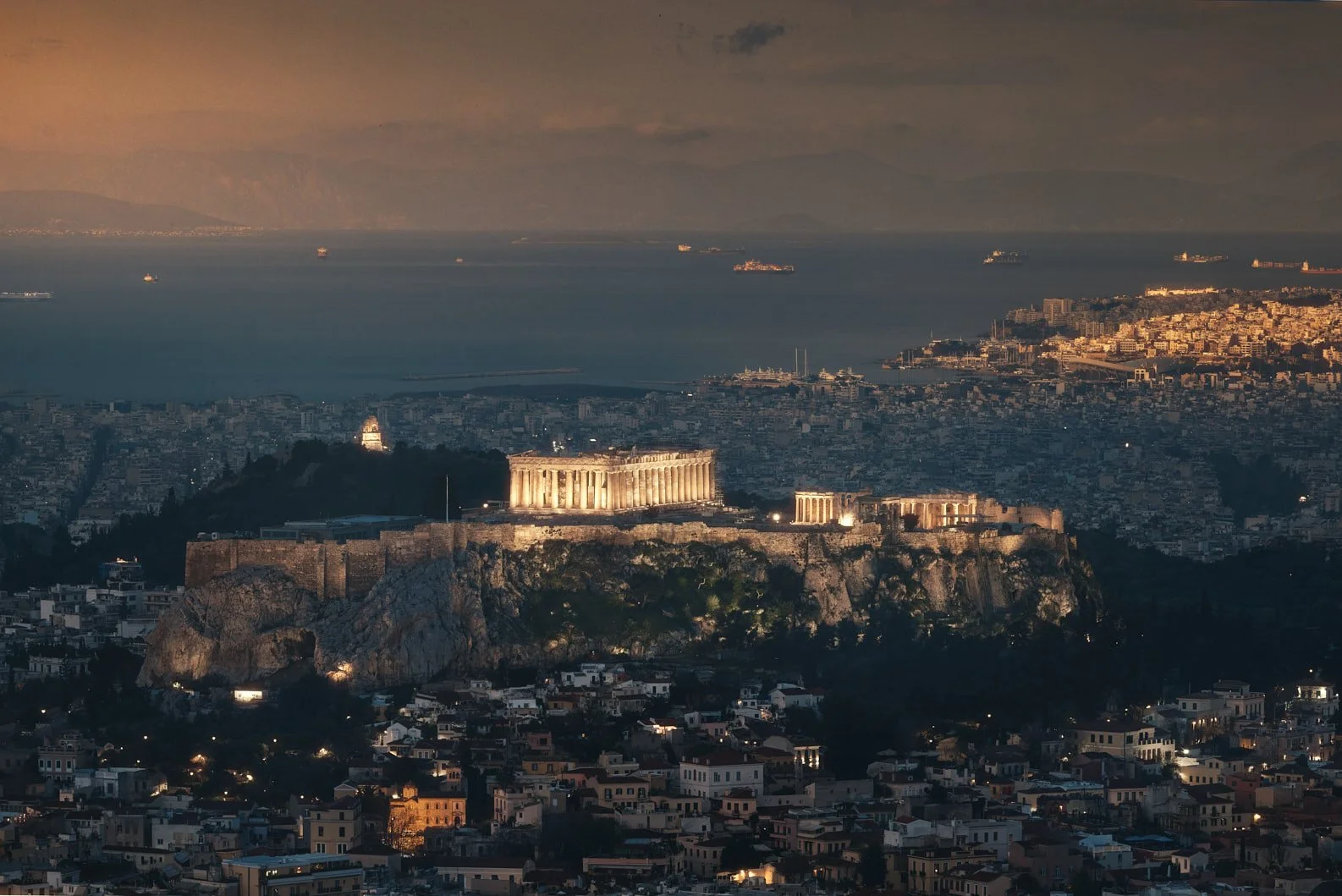 Architectural photograph of the illuminated Acropolis of Athens at golden hour, showcasing the Parthenon and surrounding ancient structures perched on a rocky hilltop above the city, with the Aegean Sea and distant mountains in the background. Captur