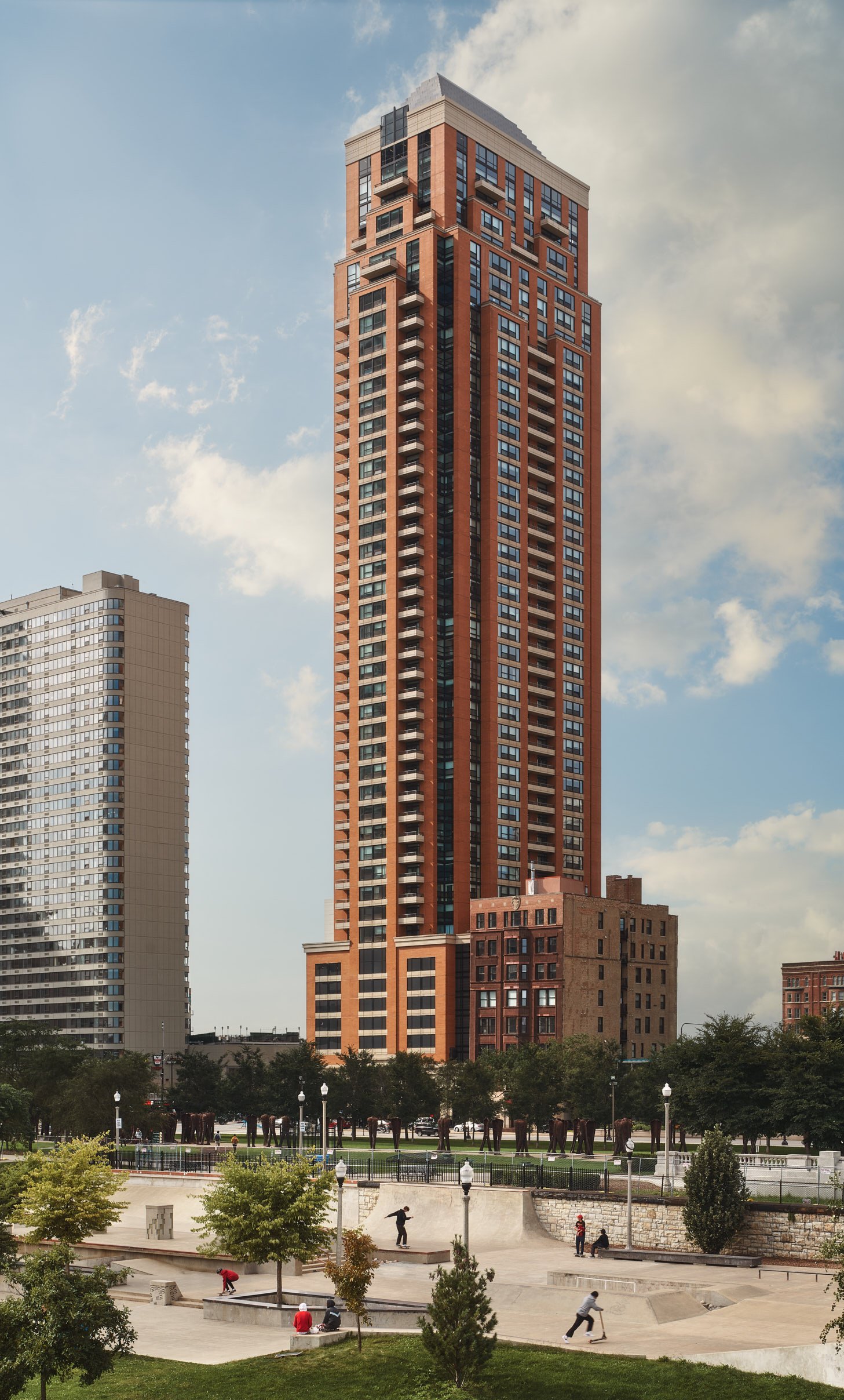 Architectural photograph of The Colombian, a red-and-beige high-rise residential skyscraper in Chicago, towering over a lively skatepark below. The image captures a mix of urban design and community activity under a partly cloudy sky, showcasing the