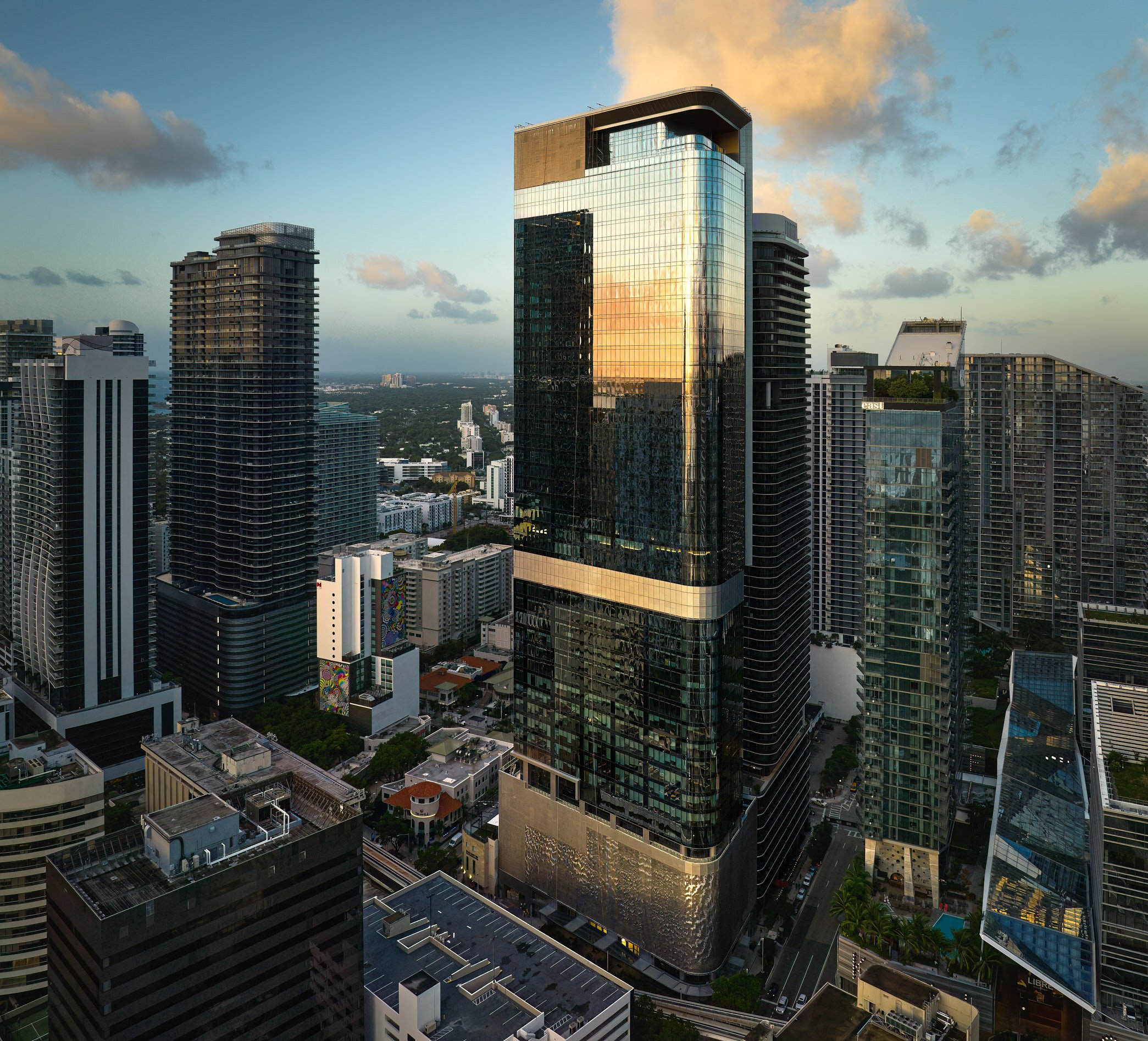 Architectural aerial photograph of the Baccarat Residences tower under development in Brickell, Miami, at golden hour. The glass facade reflects the glowing sunset while neighboring skyscrapers form a dense urban grid extending toward the horizon. Ca