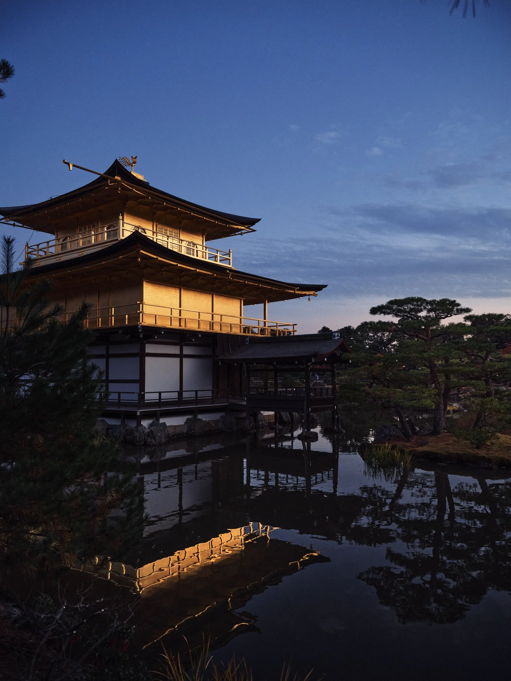 Architectural photograph of Kinkaku-ji, the Golden Pavilion in Kyoto, Japan, captured at dusk. The iconic Zen Buddhist temple glows in warm golden tones, reflecting perfectly in the still waters of the surrounding pond. Framed by lush pines and dista