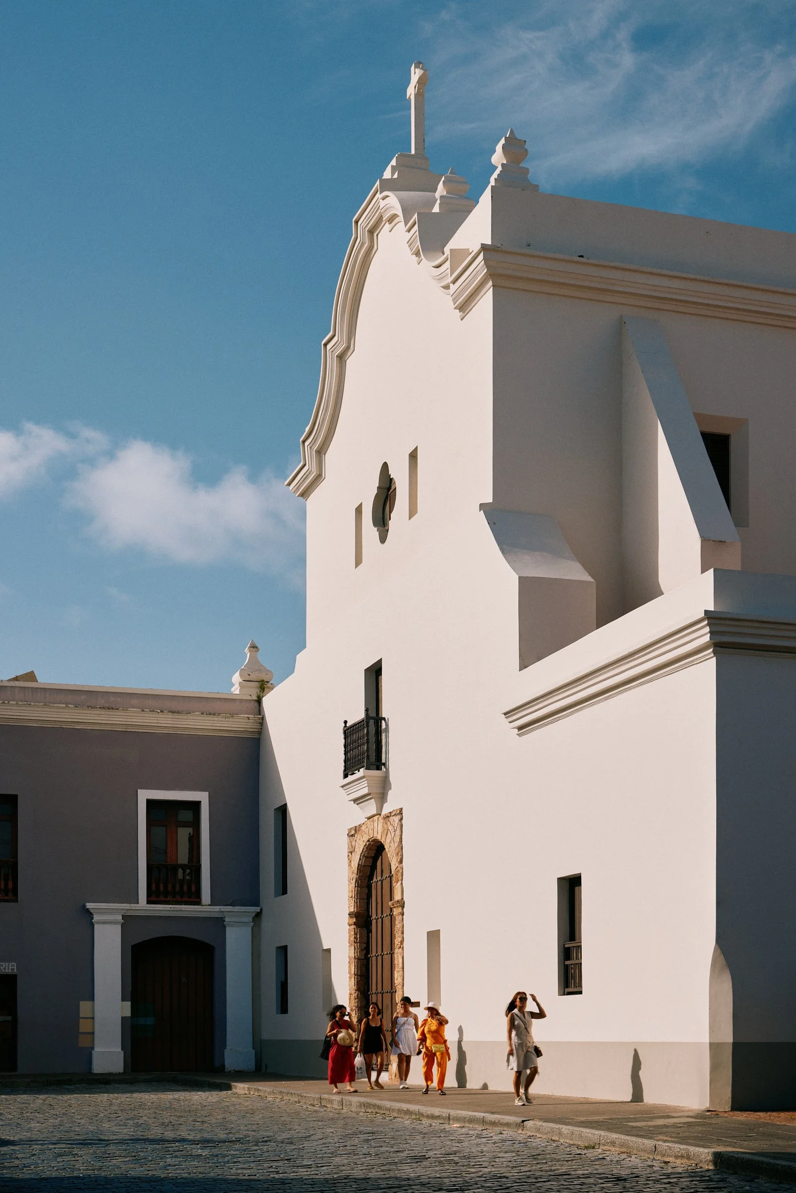 Architectural photograph of a historic whitewashed church in Old San Juan, Puerto Rico, bathed in warm afternoon light with a vivid blue sky above. A small group of people in colorful clothing walks past, adding vibrancy and a sense of local culture.