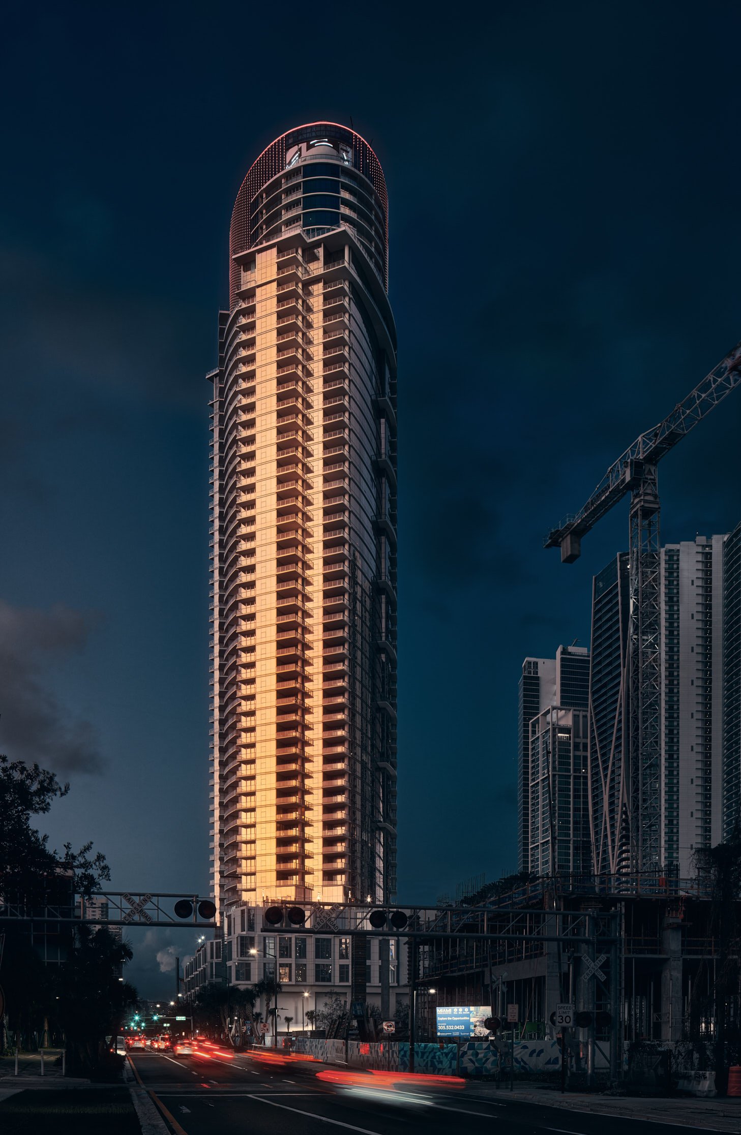 Architectural photograph of the Paramount Miami Worldcenter tower at night, its vertical glass facade glowing with warm interior lights against a moody evening sky. Light trails from passing cars and visible construction cranes add dynamic urban ener