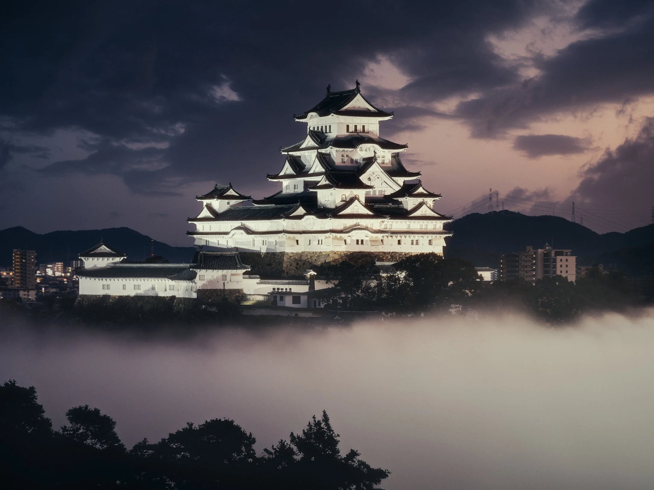 Architectural travel photograph of Himeji Castle in Japan illuminated at twilight, rising above a sea of mist with dramatic storm clouds in the background. The iconic white castle, a UNESCO World Heritage Site, contrasts against the dark mountains an