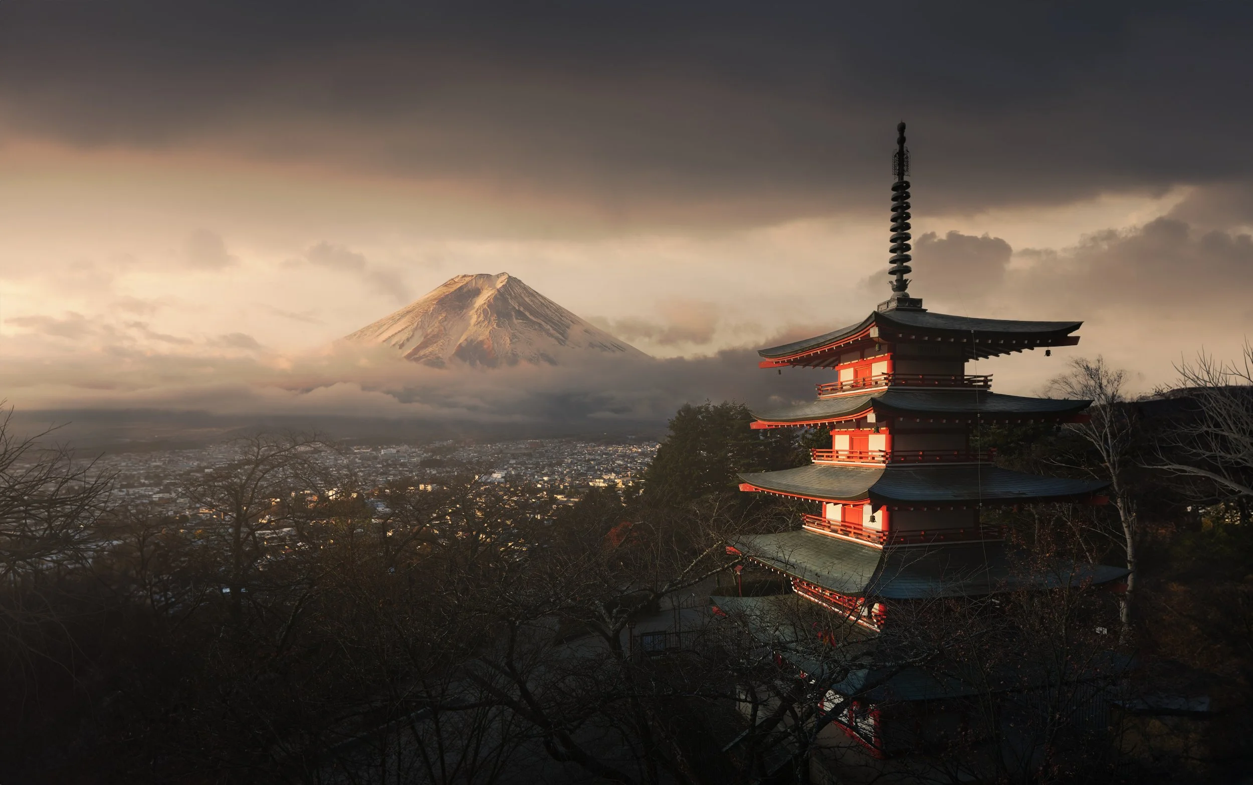 Architectural photograph of the Chureito Pagoda in Fujiyoshida, Japan, illuminated against a dramatic stormy sky with the snow-capped summit of Mount Fuji emerging through the clouds above the city below — a defining image of Japanese historical arch