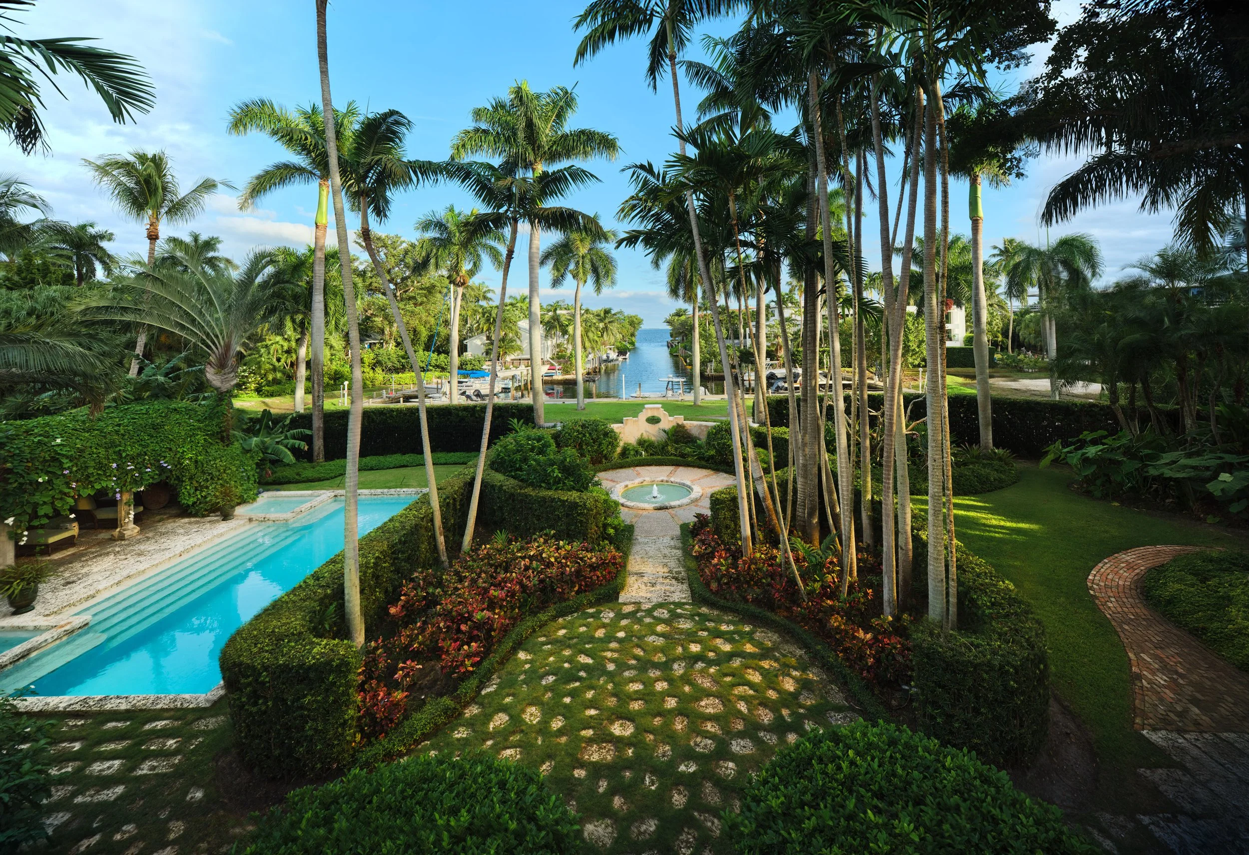 Architectural photograph of a lush tropical garden with winding stone pathways, towering palms, and dense greenery enveloping the Mediterranean Revival estate. Captured by Miami and Los Angeles-based architectural and travel photographer Christian Sa