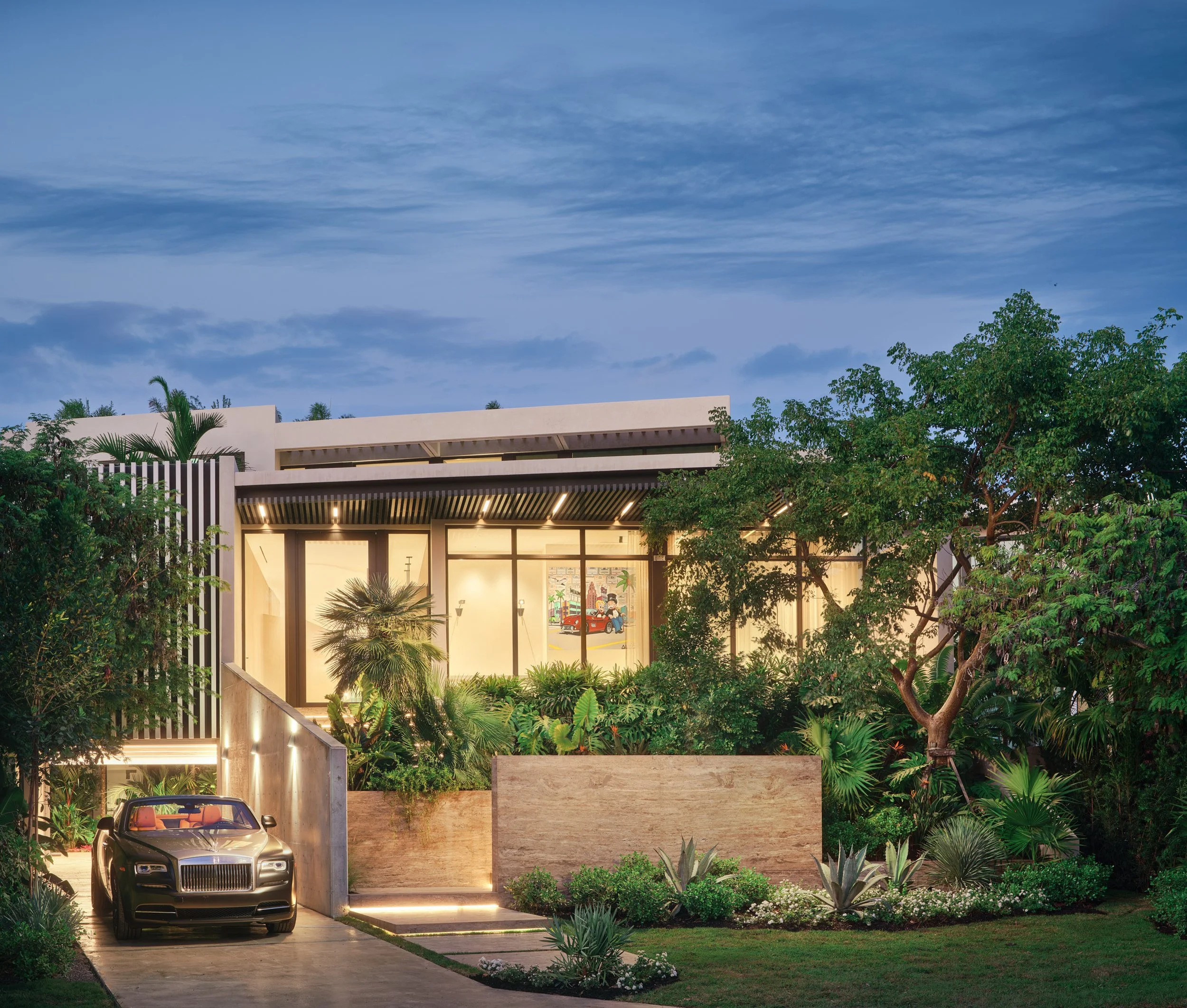 Architectural photograph of a modern Venetian Islands waterfront residence in Miami at twilight, featuring layered tropical landscaping, limestone walls, glass façade, and a luxury car arriving through the driveway entrance. Captured by Miami and Los