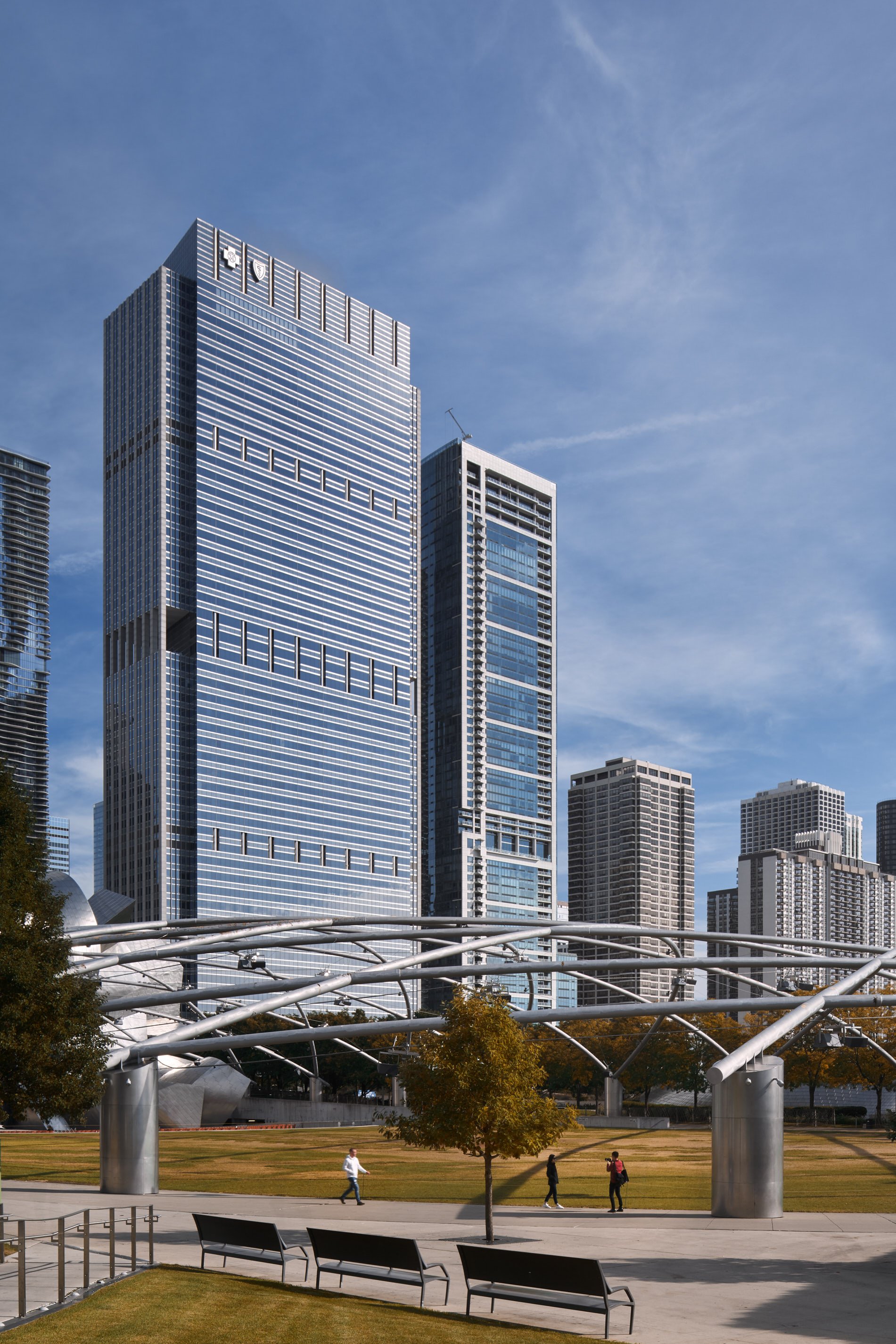 Architectural photograph of the Blue Cross Blue Shield Tower in downtown Chicago, captured from Millennium Park on a clear autumn day. Sleek glass façades and clean lines define the modern skyscraper, with the Pritzker Pavilion’s sculptural trellis i