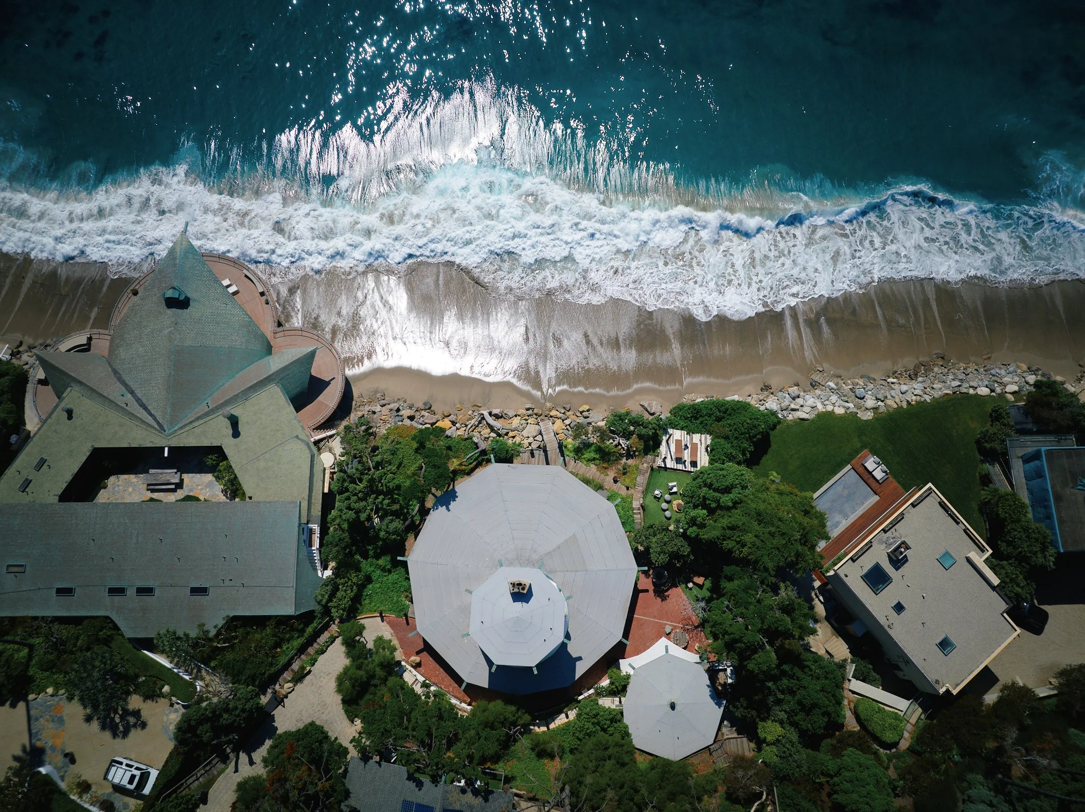 Aerial architectural photograph of Harry Gesner’s Sandcastle House in Malibu, California, situated directly on the shoreline. The image reveals the octagonal plan, sculptural tower, and lush landscaping nestled between modern neighbors. Aerial perspe